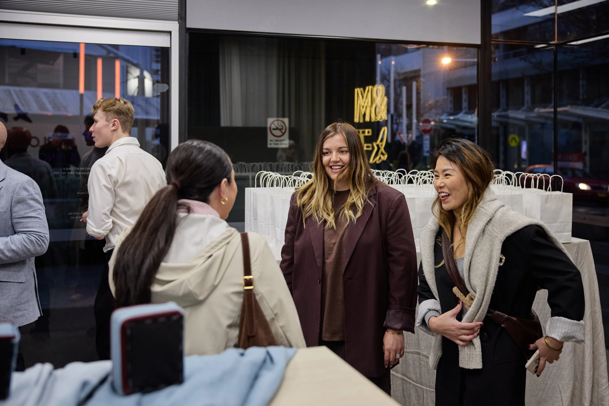 Group of people engaging in conversation inside a store or event space with gift bags in the background. It is evening outside.