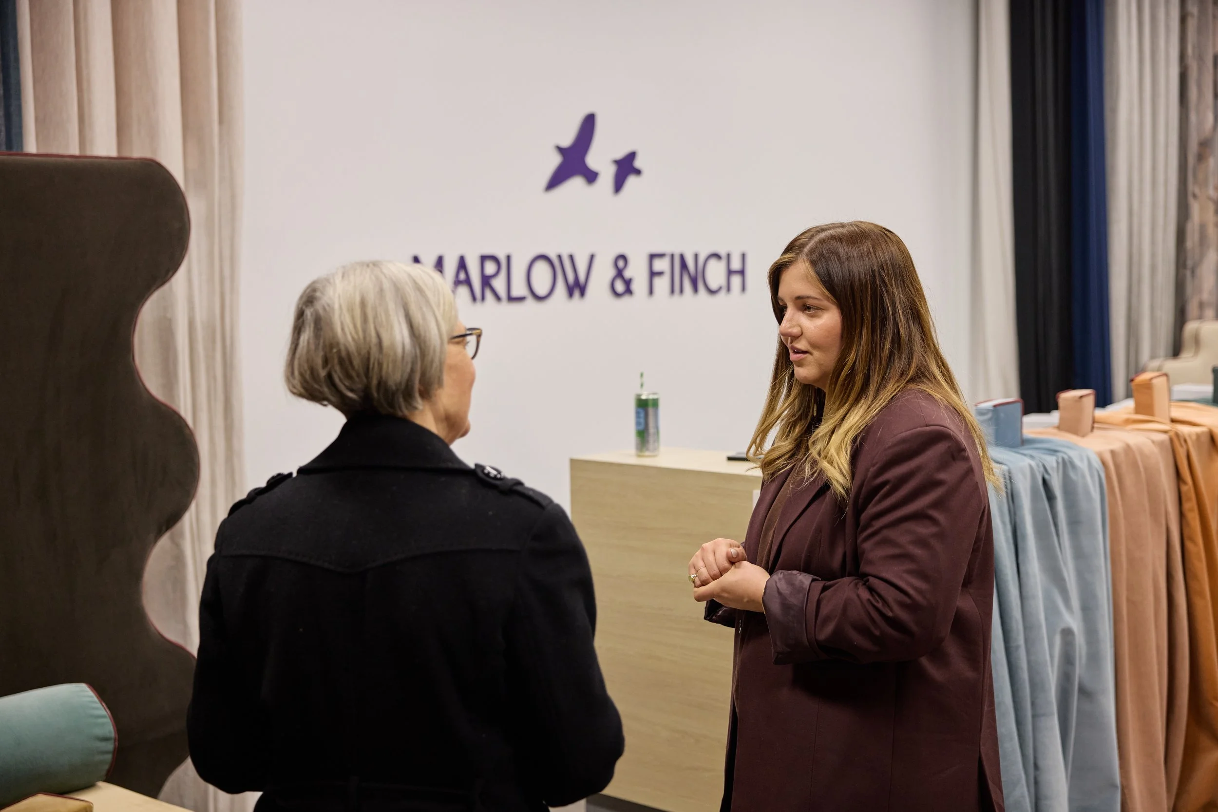 Two women having a conversation in an indoor setting, with the words 'Marlow & Finch' and purple bird logos on a white wall in the background.