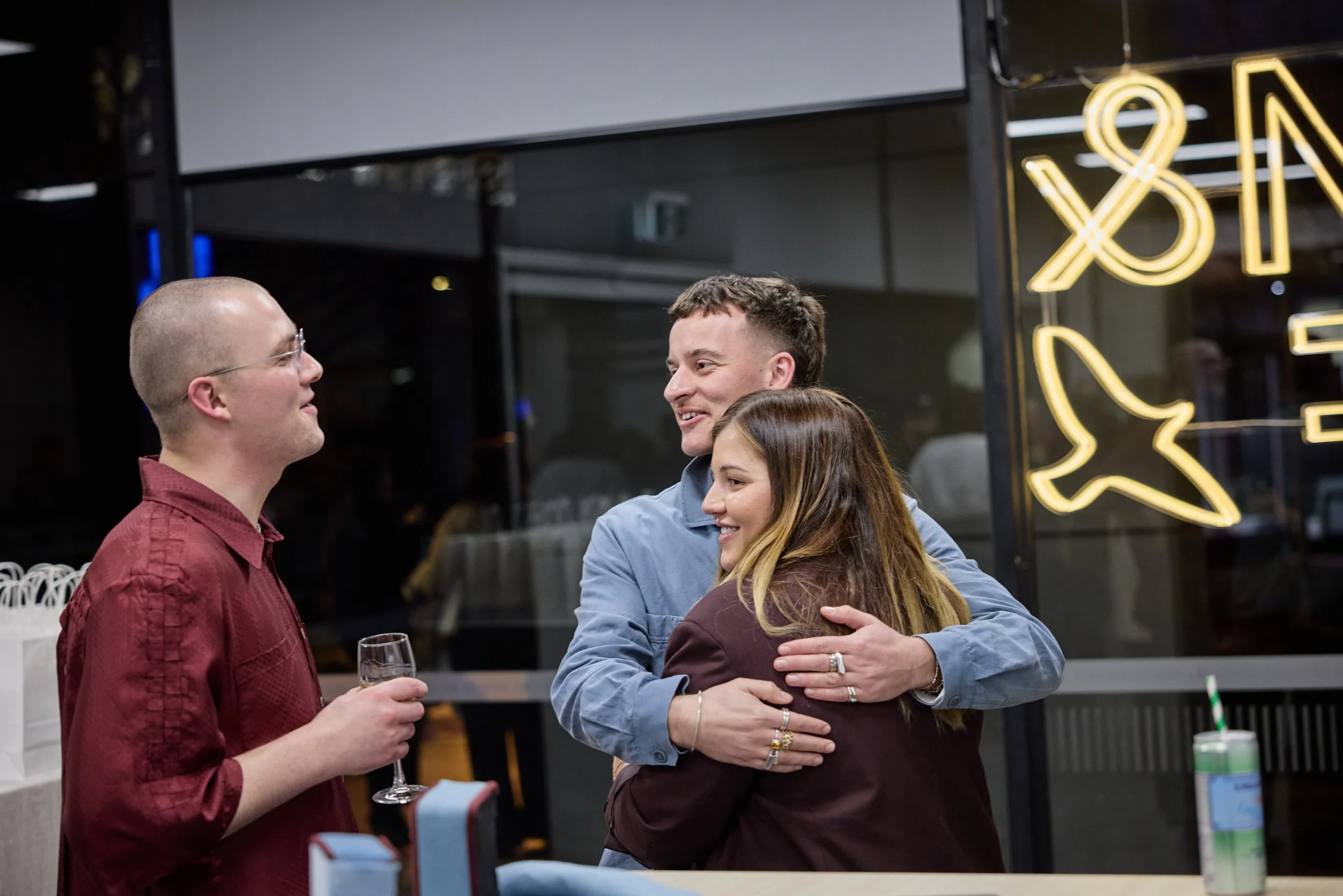 Three people at a social gathering, with one man in a red shirt holding a glass, hugging another man and woman who are smiling.