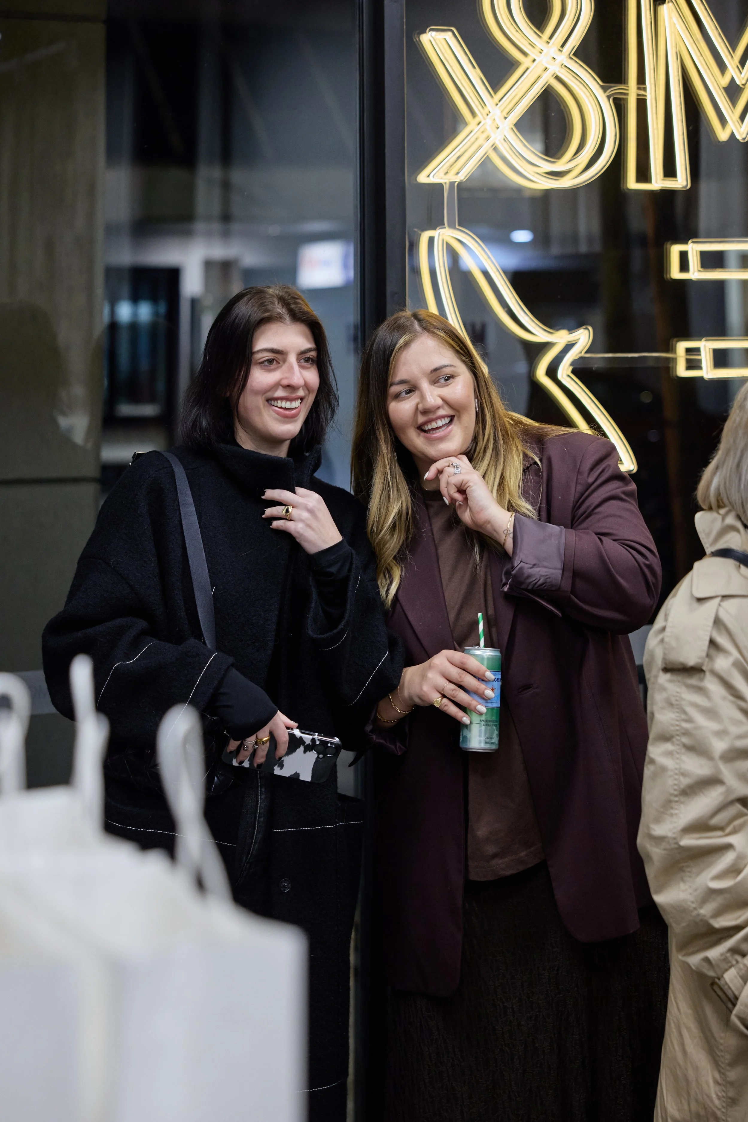 Two women standing outside a store, smiling and chatting. One woman has black hair and is wearing a black coat, holding a phone. The other woman has blonde hair, wearing a brown coat, holding a can with a green straw. Neon signs are visible in the ba