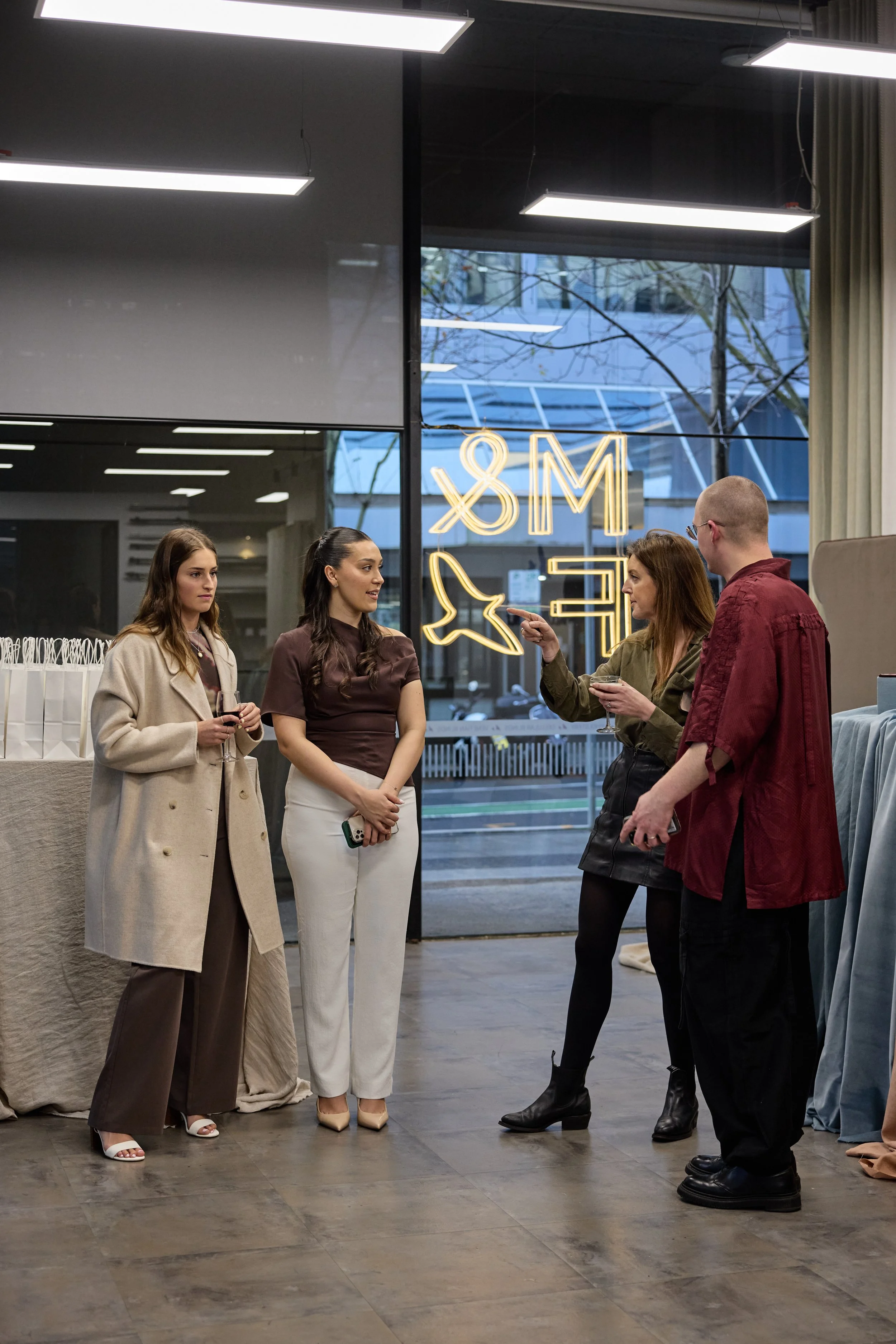 Four people in a retail shop or gallery having a conversation. Two women are holding glasses of wine, one woman and one man are talking, with the woman pointing at the man. Behind them are window with neon signs and a street view.