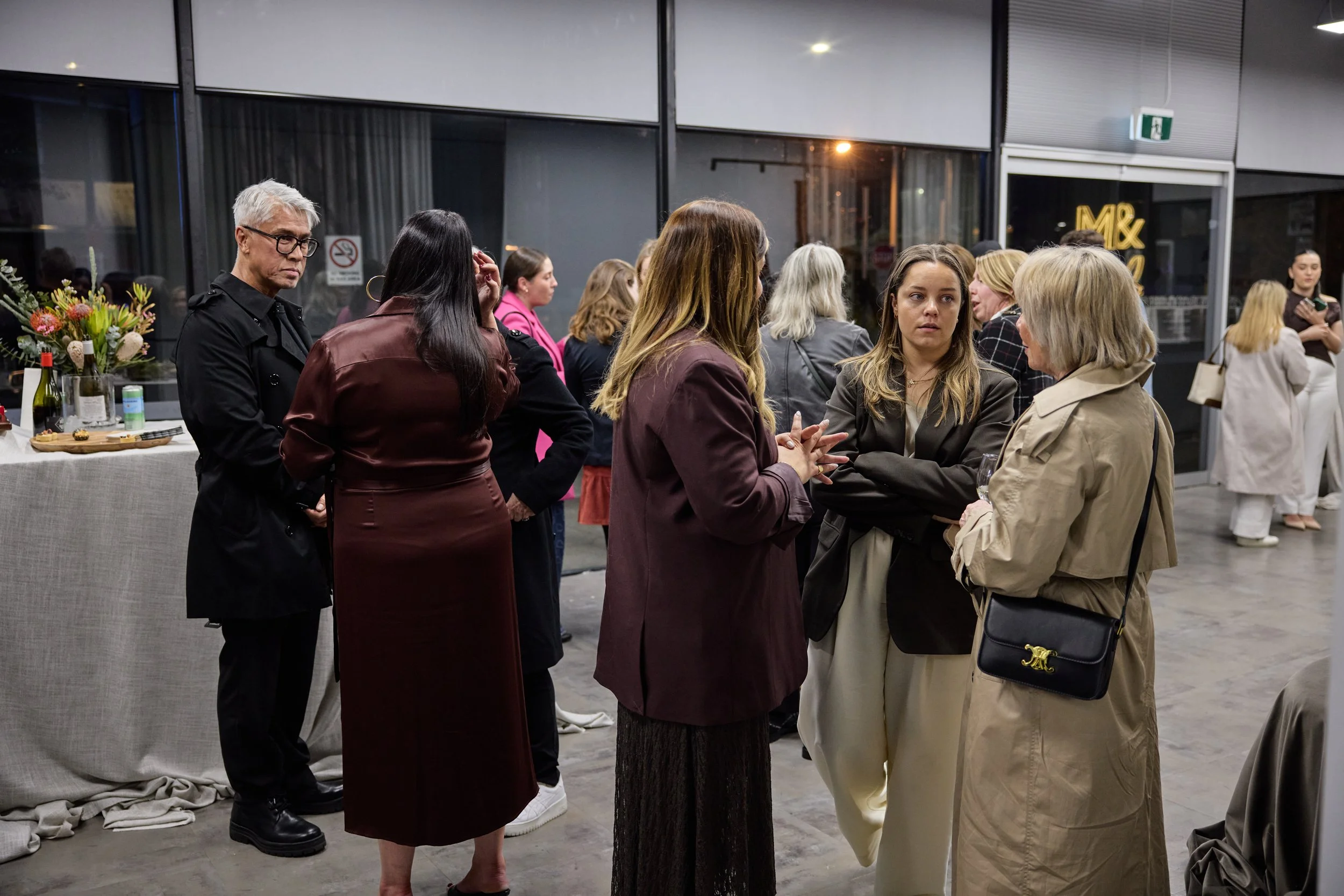 People socializing at an indoor event, some holding wine glasses, in front of large windows with city lights visible outside.