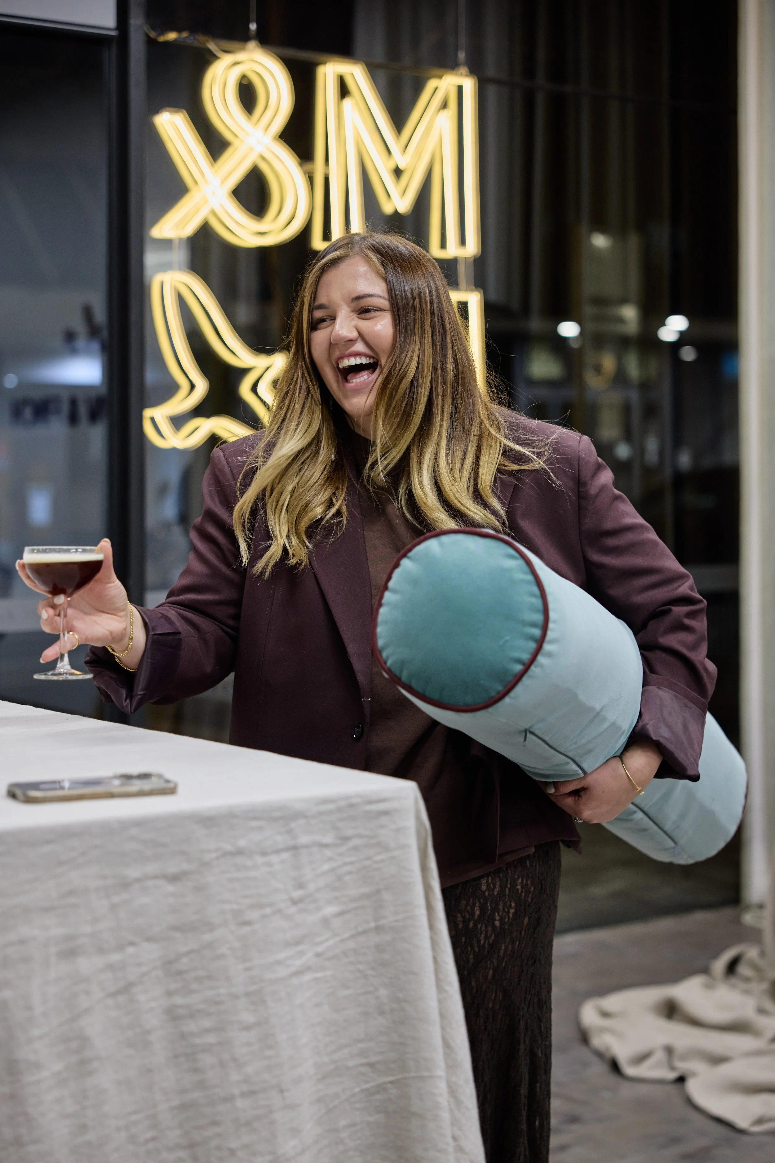 A woman with long, wavy blonde hair, smiling and laughing, holding a glass of dark beverage in one hand and a rolled-up pillow in the other, standing in a room with a glowing neon sign in the background that reads '8 M' and a lemon graphic.