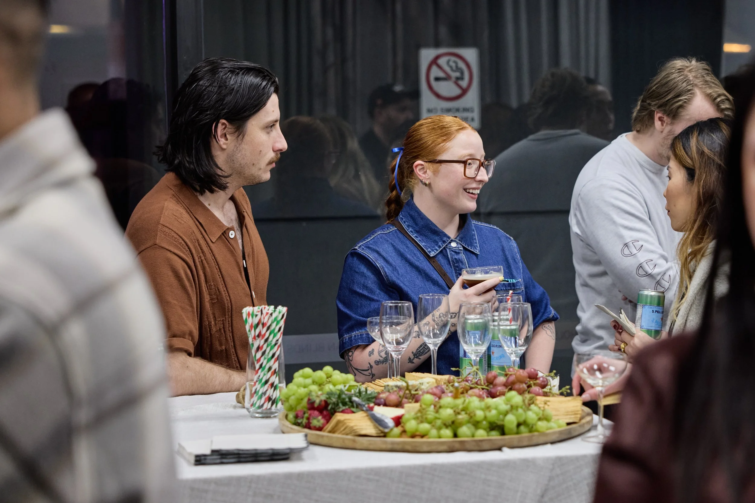 A group of people at a social gathering, with a woman in a blue denim shirt holding a drink, surrounded by glasses and a tray of grapes and snacks on a table.