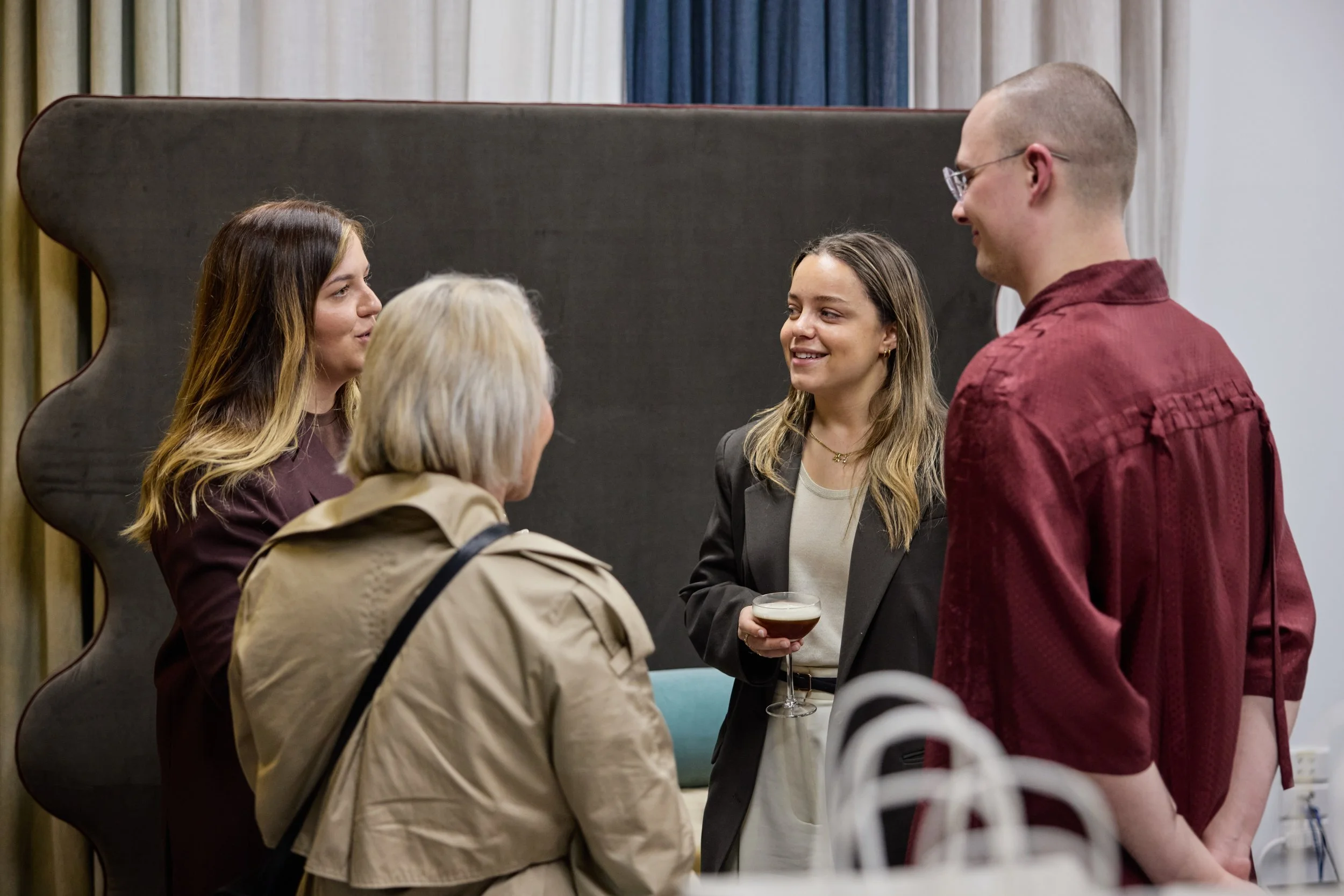 Group of five people talking at social gathering, one person holding a drink, in an indoor setting with curtains and a dark backdrop.