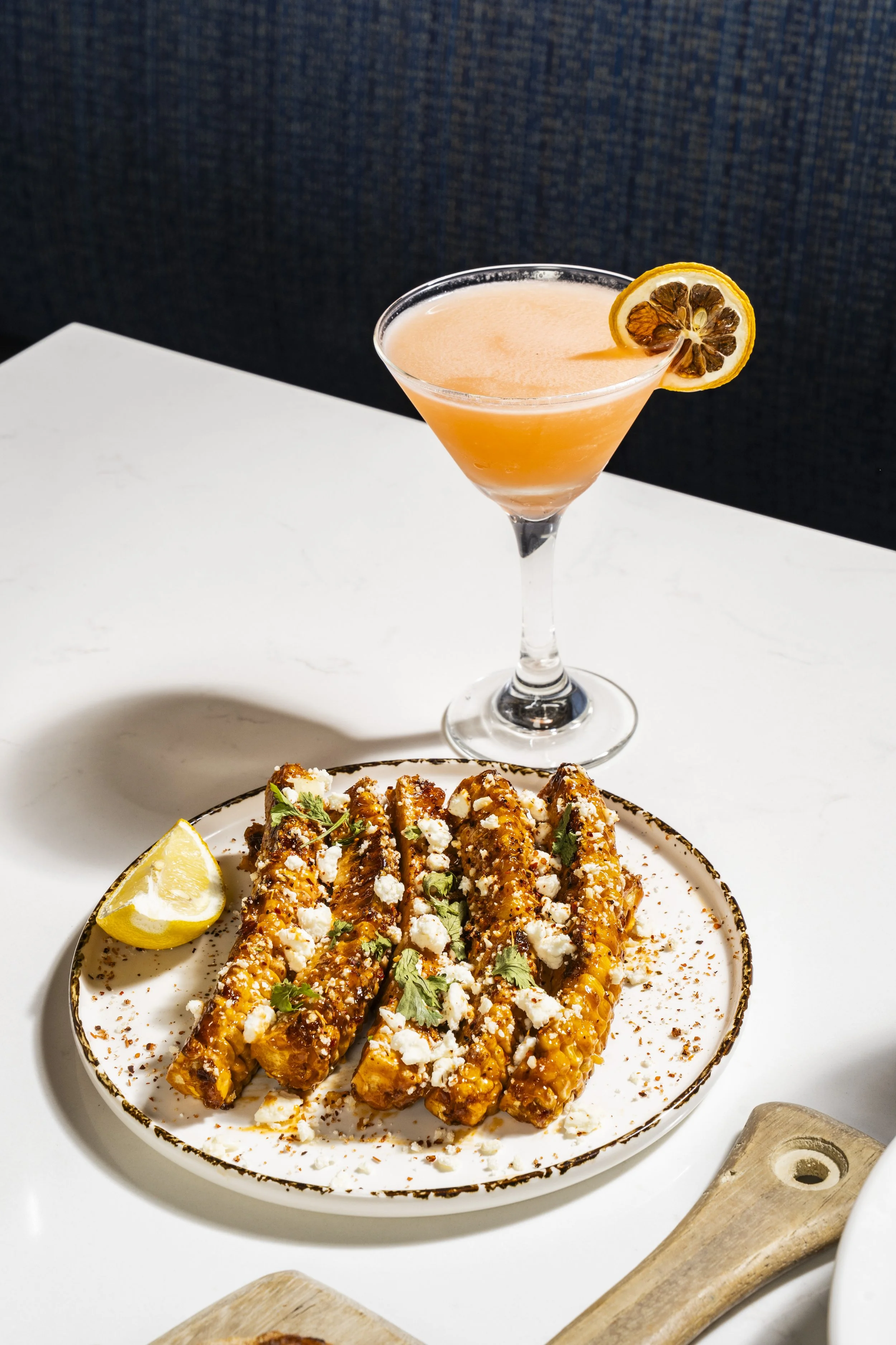 Plate of Mexican street corn topped with cheese and herbs, lemon wedge on side, sour cream in background, cocktail with lemon slice garnishment on a white table.