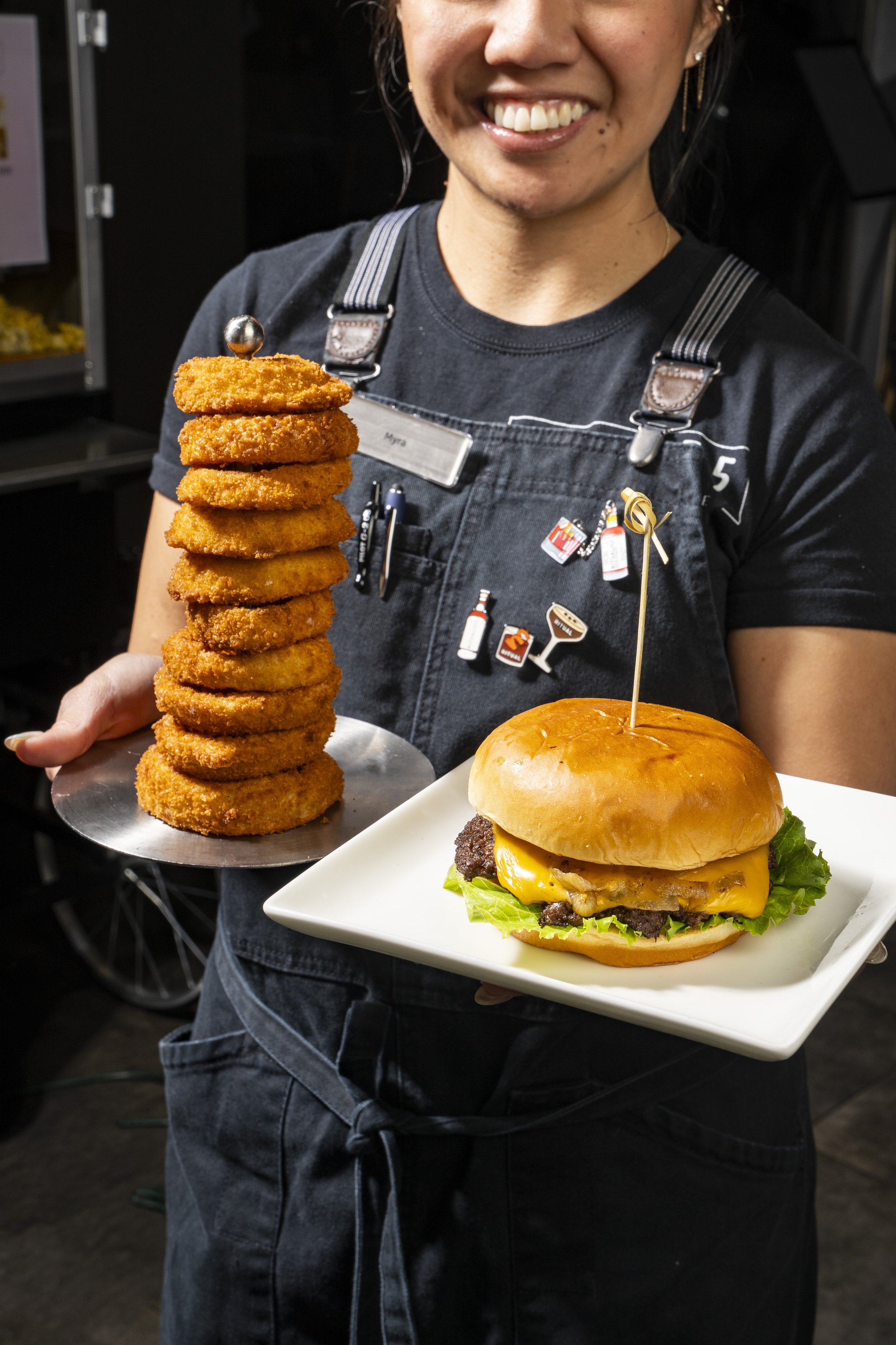 A woman smiling while holding a tall stack of breaded onion rings in her right hand and a plate with a cheeseburger on it in her left hand. The woman is wearing a black T-shirt with a gray apron, and there are various pins and badges on the apron. The background is dark and blurry.