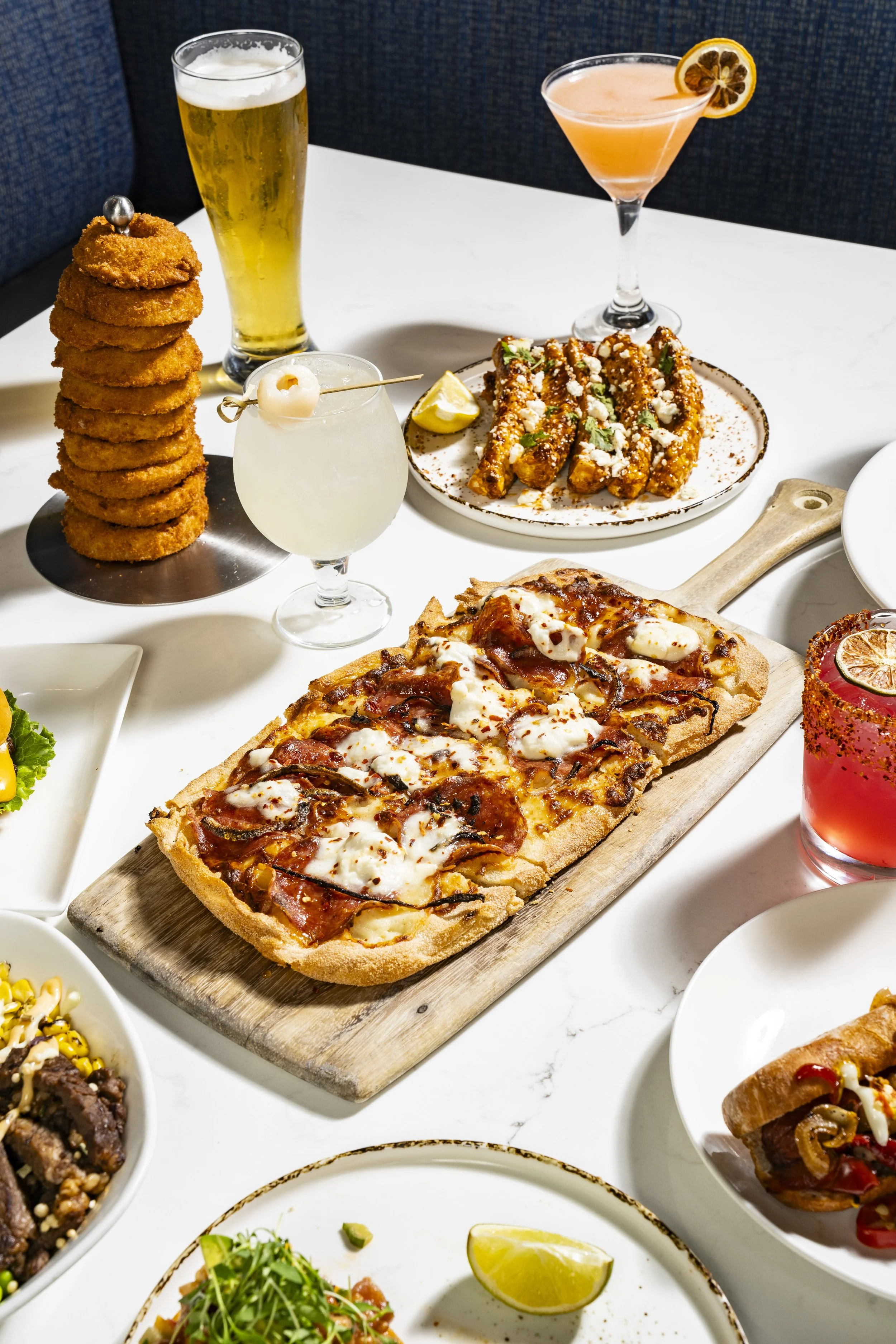 Assorted dishes including pizza, fried onion rings, various cocktails, salads, and other appetizers on a white table.
