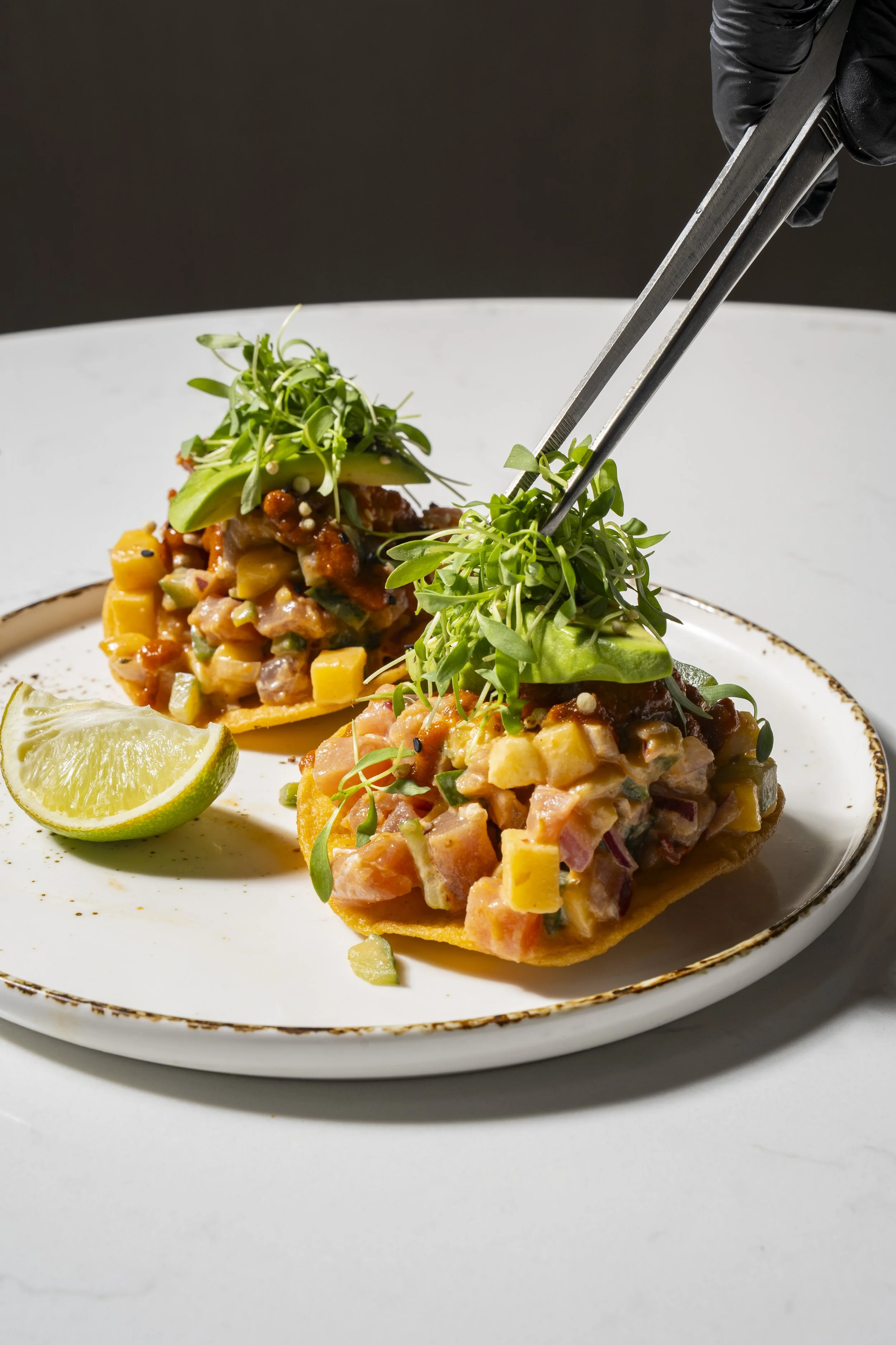 Three Asian-style sushi tacos topped with microgreens and a wedge of lime on a white plate.