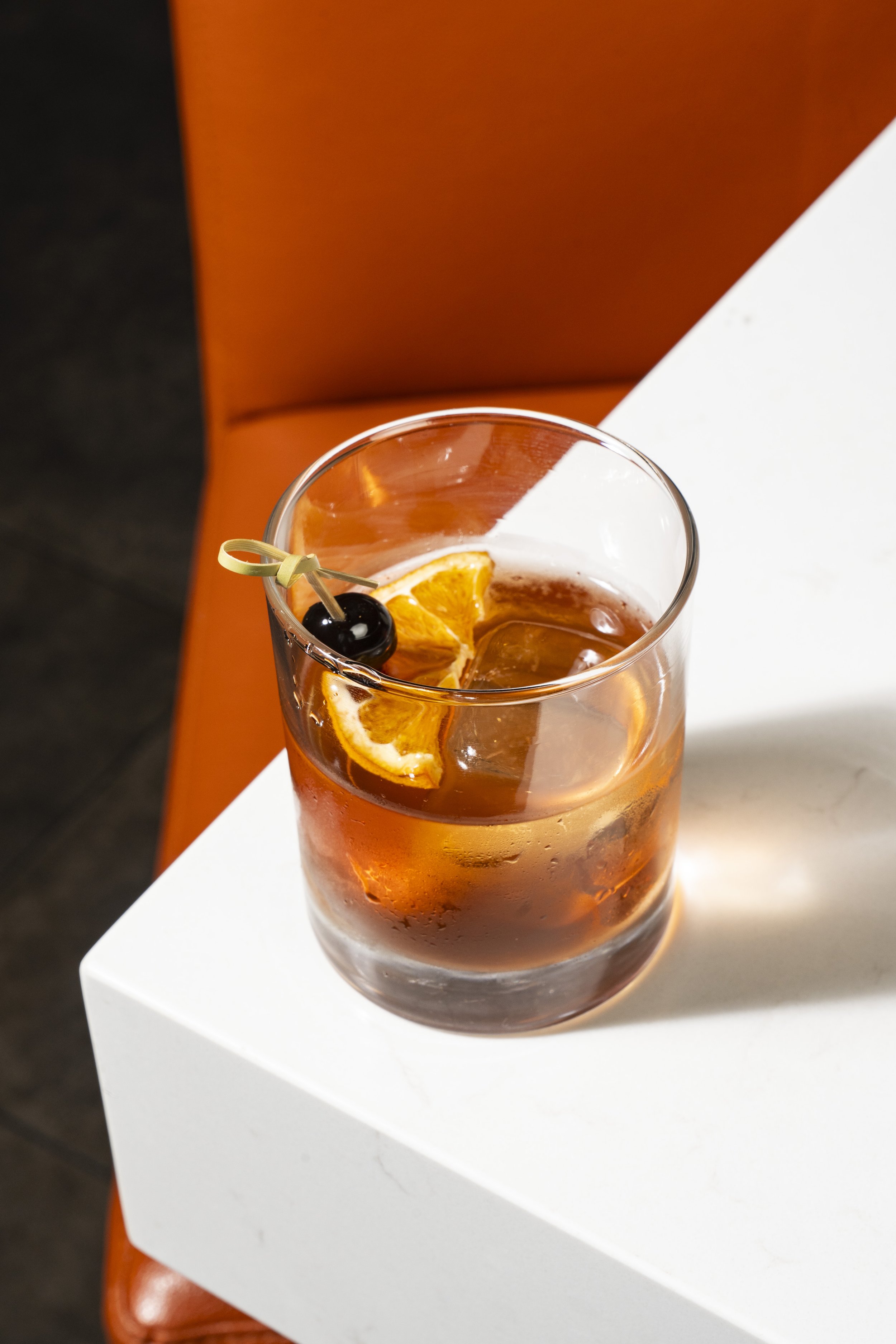 A glass of iced beverage with an orange slice and a black cherry garnish on a white surface, with an orange background. Monterey Park cocktails