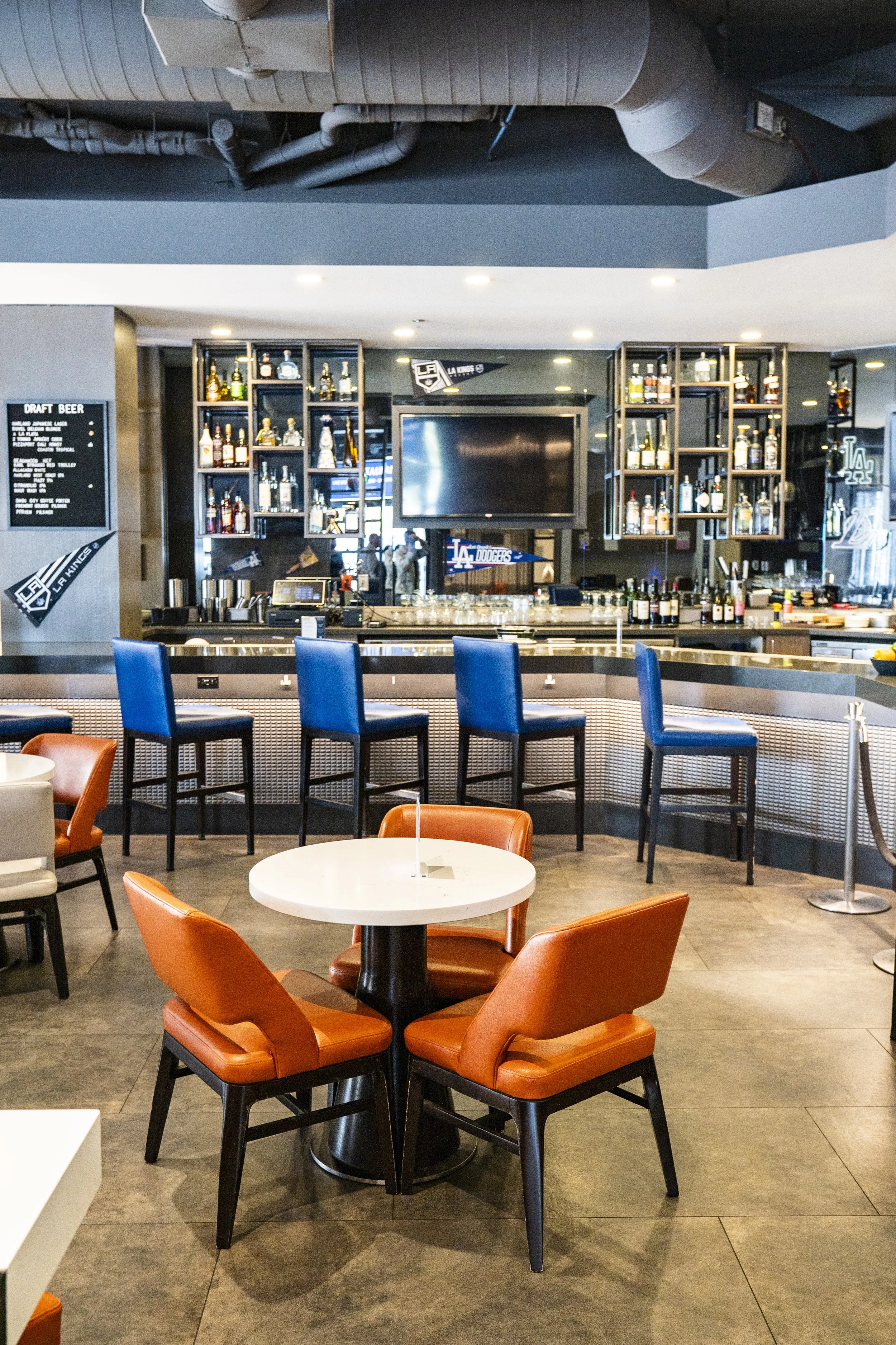 Interior of a bar or restaurant featuring a bar counter with orange and blue chairs, a wall-mounted flat screen TV, and shelves with liquor bottles behind the bar. There are additional seating areas with round tables and chairs.