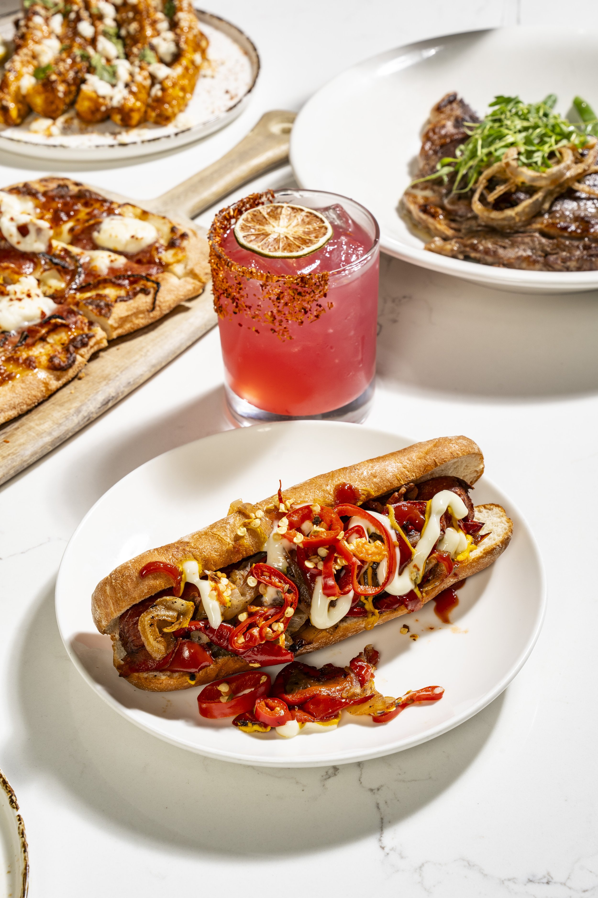 A hot dog topped with sliced red peppers, onions, and mayonnaise in a white bowl, with various dishes including a pizza slice, a plate of grilled meat, a plate of fried chicken, and a pink cocktail with a lime slice in the background. Craft Cocktails