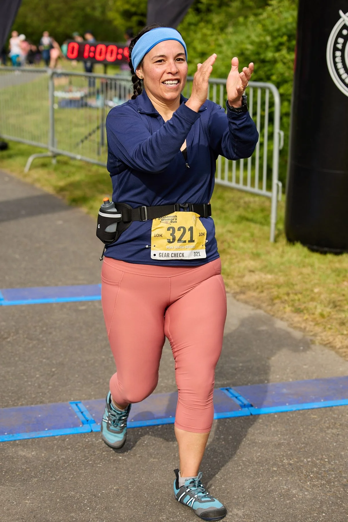 Female runner crossing the finish line at a race, smiling, wearing a blue jacket, pink pants, and a blue headband. Race bib number 321 is visible, with a water bottle attached to her waist. The background shows a digital timer, a black inflatable arch, and a metal fence with spectators.