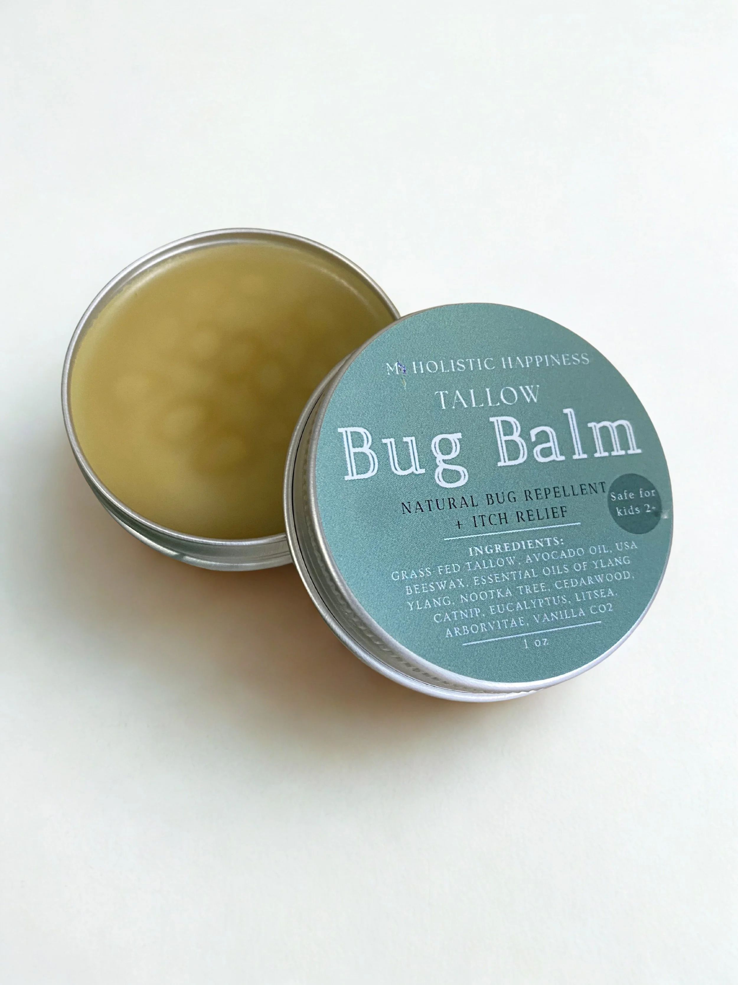 A metal tin filled with bug balm stands on a white surface with the lid labeled tallow bug balm by My Holistic Happiness