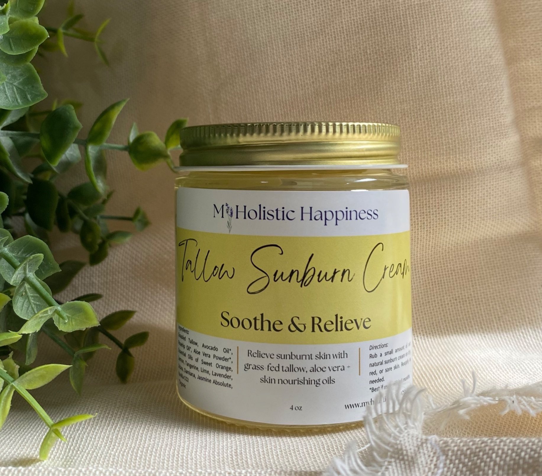 The sun shines on a glass jar labeled Tallow Sunburn Cream by My Holistic Happiness stands on a cream colored fabric. A green eucalyptus plant stands in the corner.