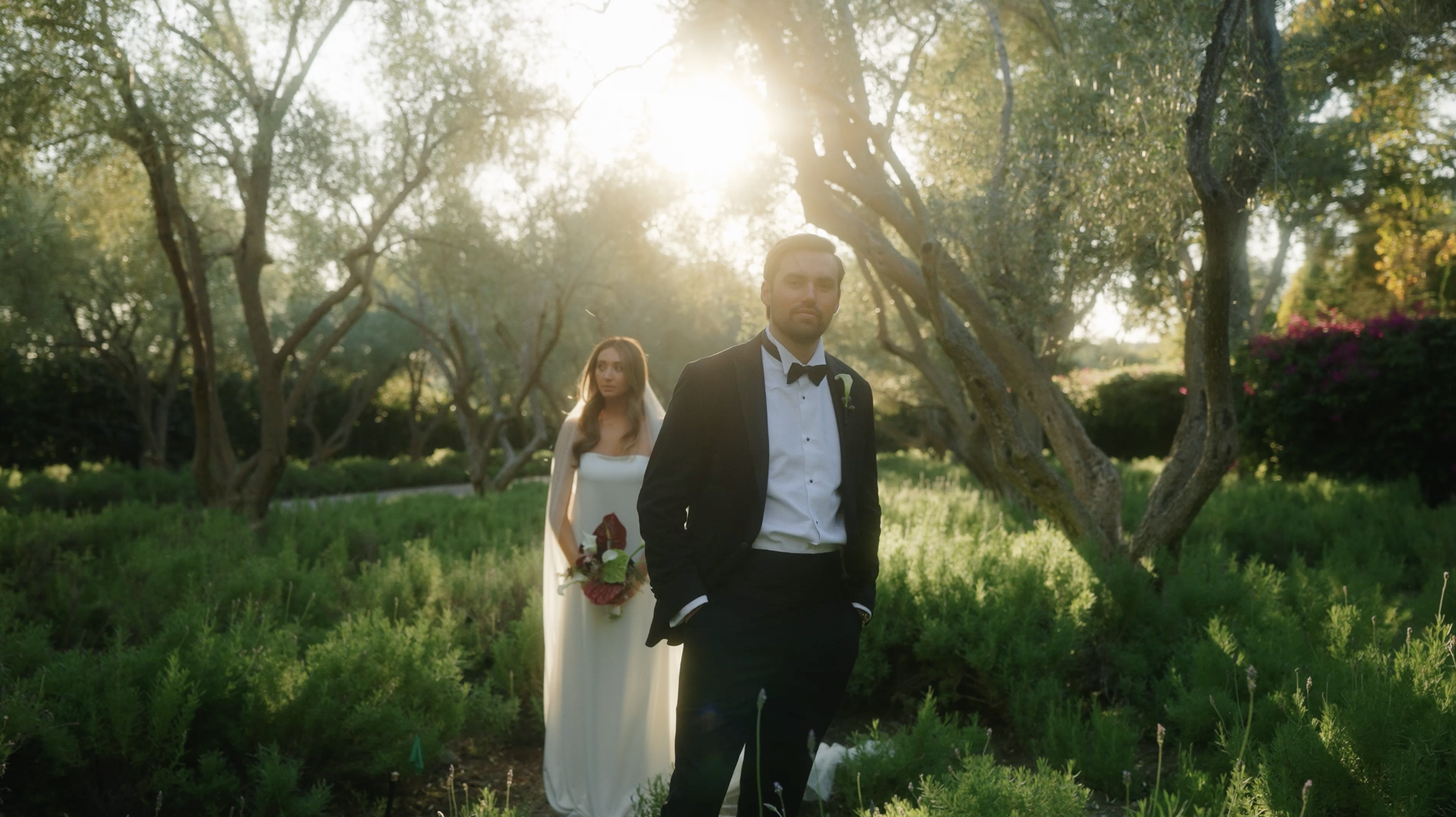 A groom in a black tuxedo with a bow tie standing in the foreground with his hands in his pockets, and a bride in a white wedding dress holding a bouquet standing behind him in an outdoor garden during sunset.