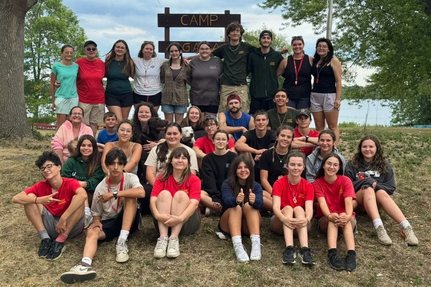 Group of young people and a few adults posing in front of a sign that says 'Camp' outdoors near a lake, with trees and cloudy sky in the background.