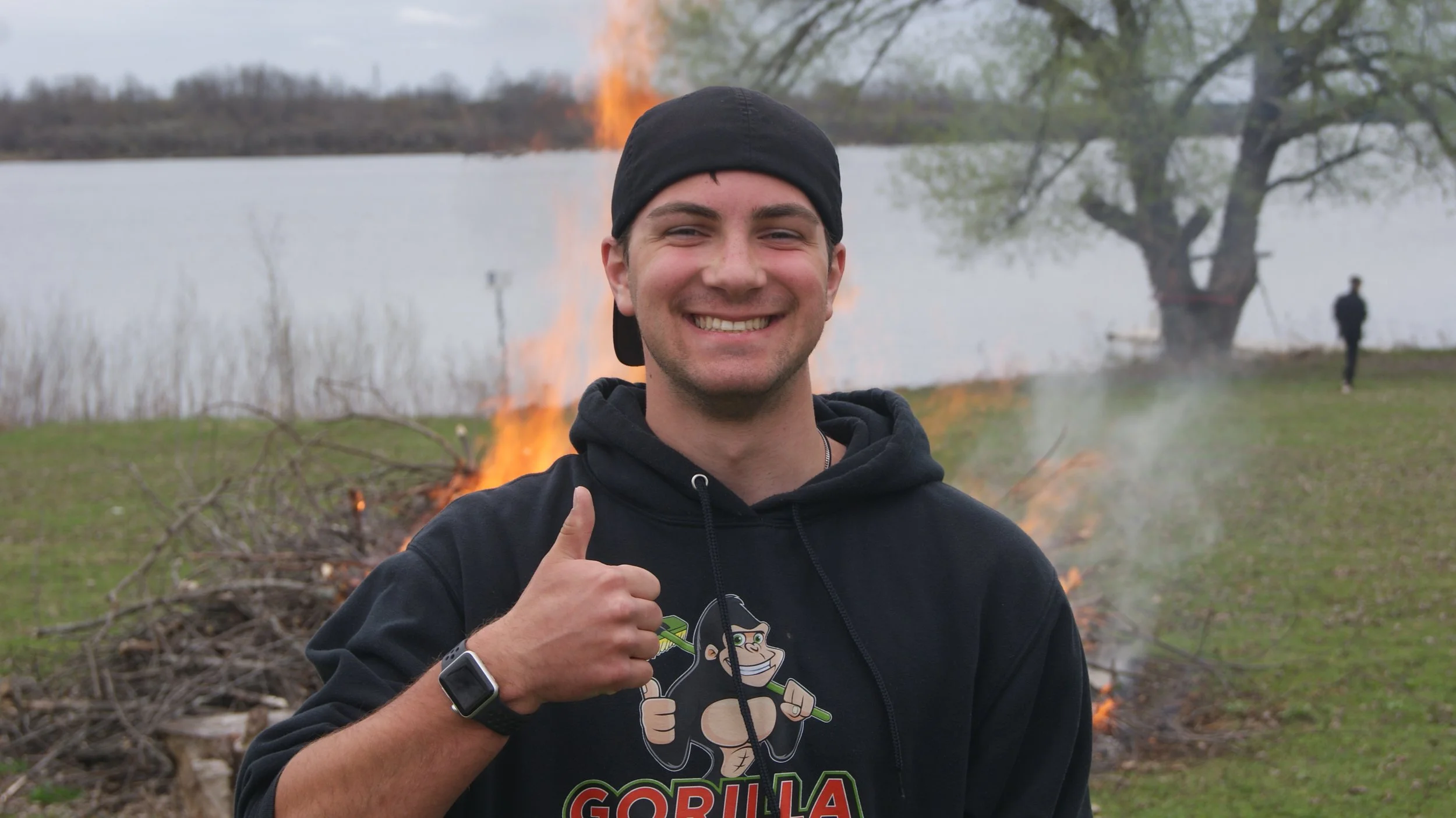 Young man smiling and giving a thumbs-up in front of a small fire near a lake with a tree and a person in the background.