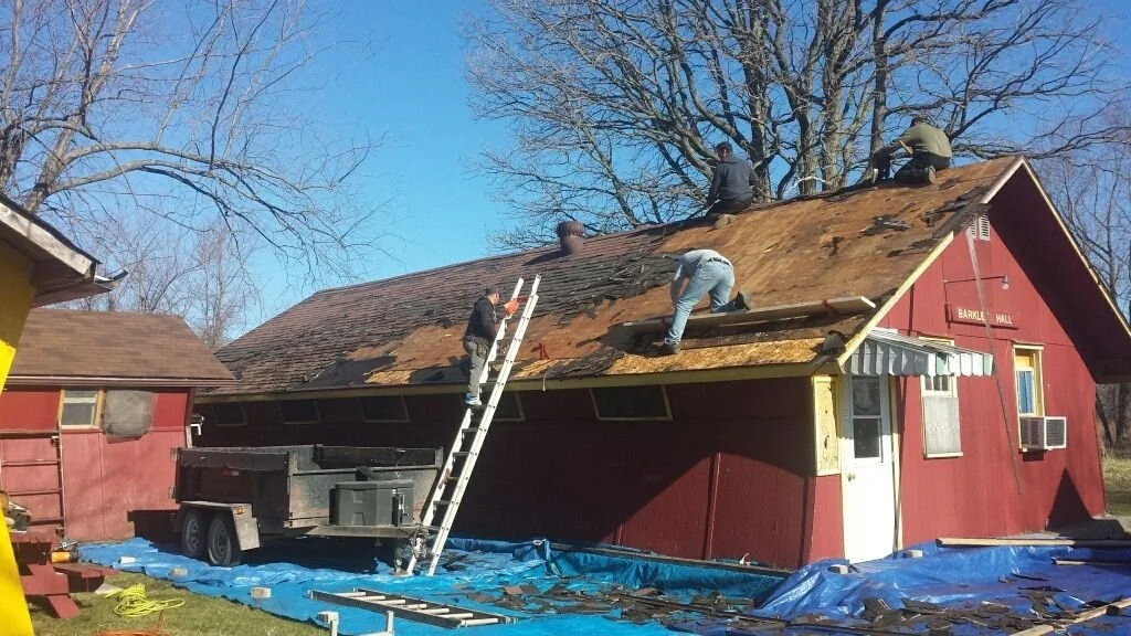 People working on a roof renovation, replacing or repairing roof shingles on a red barn-style building in a rural setting, with a trailer parked nearby and trees in the background.