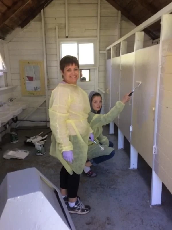 Two women in yellow protective gowns and purple gloves painting bathroom stalls in a rustic, unfinished building.