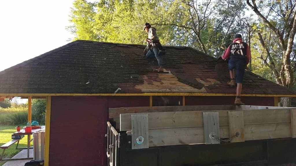 Two children working on replacing shingles on the roof of a small house or shed in a backyard.