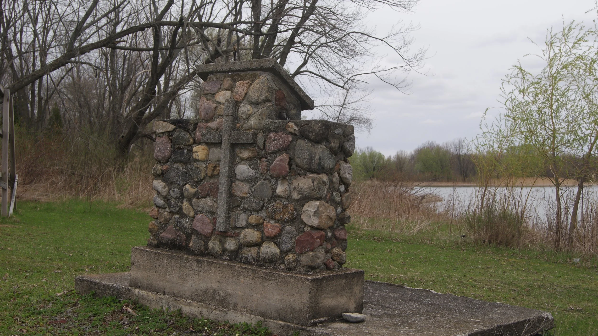 A stone monument or memorial structure near a body of water, with a concrete base and a cross carved into the stones, surrounded by grass, trees, and bushes in a natural outdoor setting.