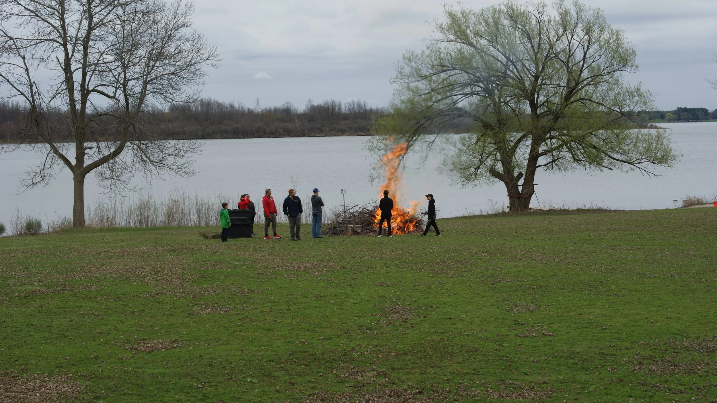 A group of people standing near a small controlled fire by a lake, with trees and cloudy skies in the background.