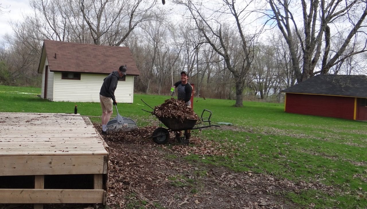 Two people working outdoors on a yard, one raking leaves and the other with a wheelbarrow filled with leaves, in a yard with trees and buildings in the background.