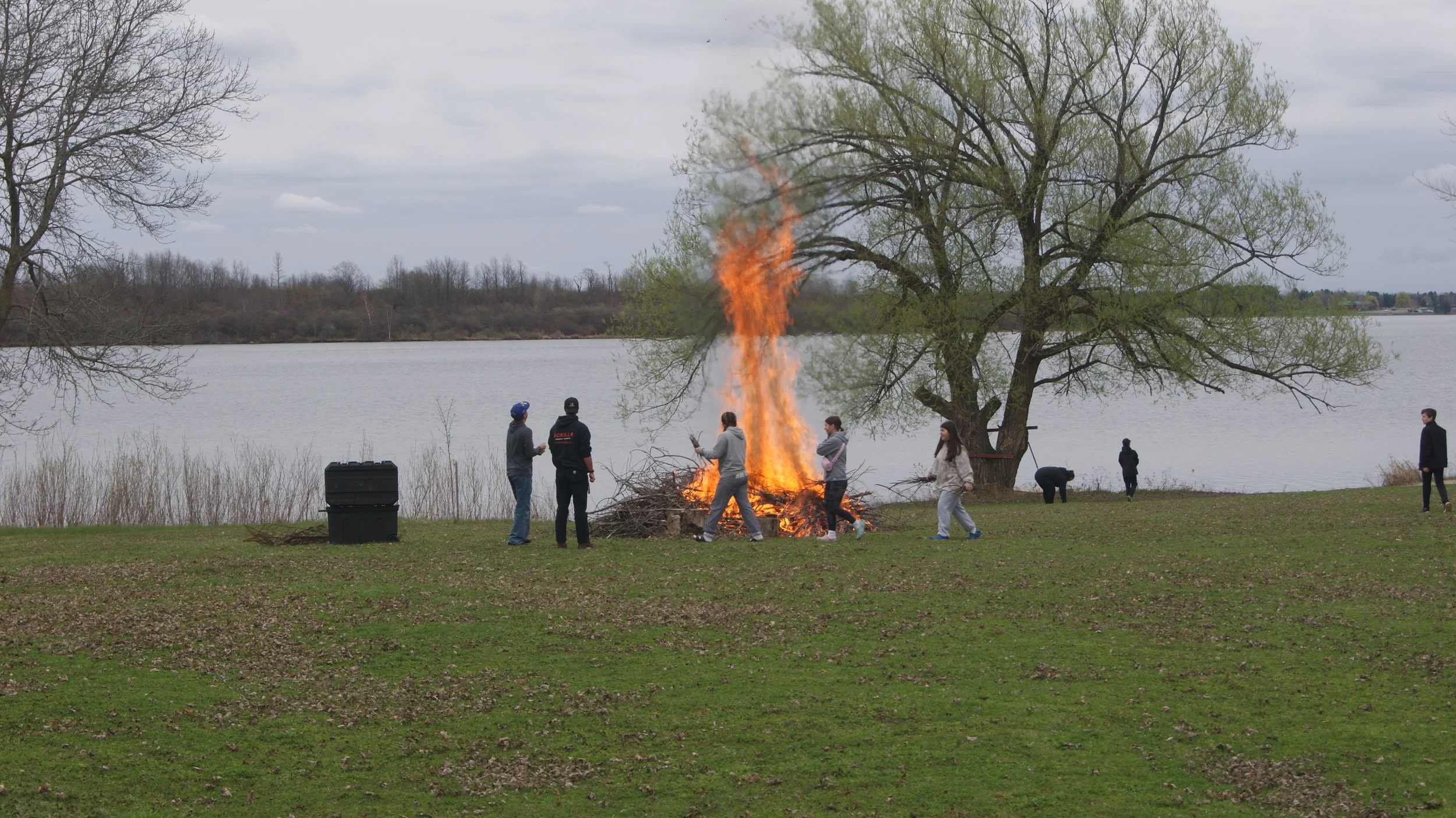 People gathered around a campfire near a lake with trees in the background, some are standing and some are bending down or walking, during an overcast day.