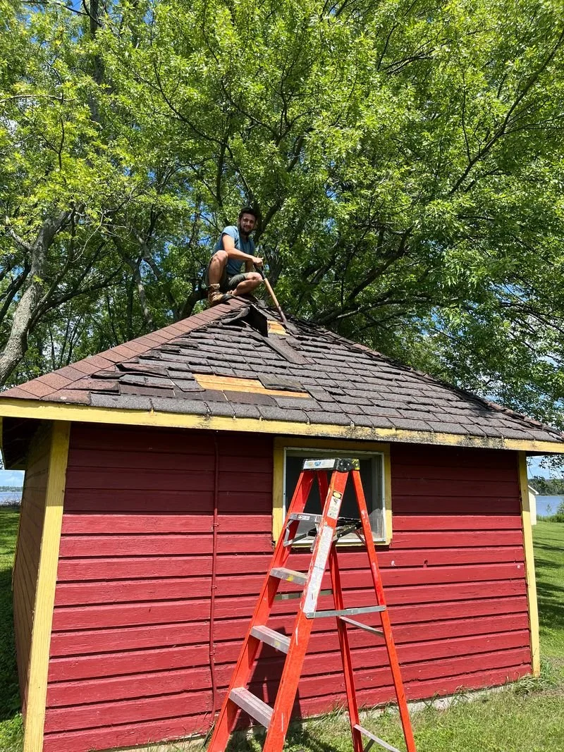 A man crouching on the roof of a small red wooden house, working on the shingles, with a stance that suggests he is repairs or inspecting the roof. A red ladder is leaning against the house below the window, and there are green trees in the backgroun