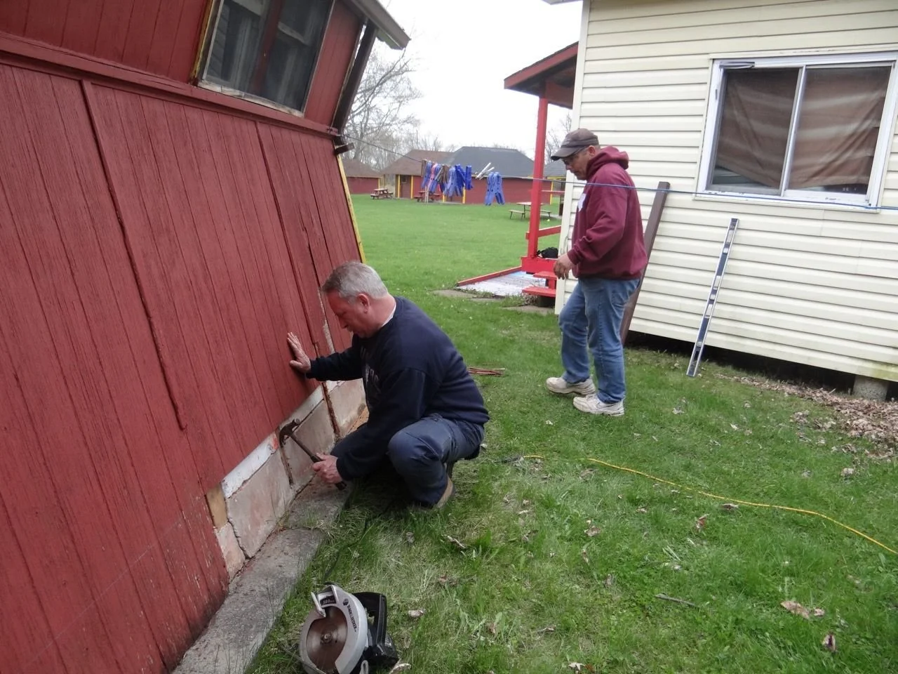 Two men working outside a house, one kneeling and hammering at the base of a red wooden wall, the other standing nearby in a burgundy hoodie and jeans, observing. The scene takes place on a grassy yard, with a white house and outbuildings in the back