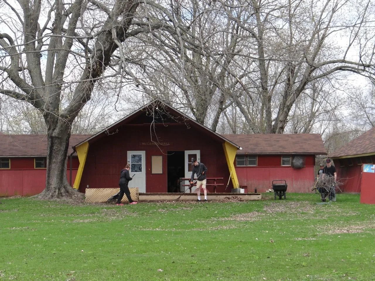 People working on a red building with yellow accents, cleaning up fallen leaves in front of it. Large trees surround the building, and the grass in the foreground is green.