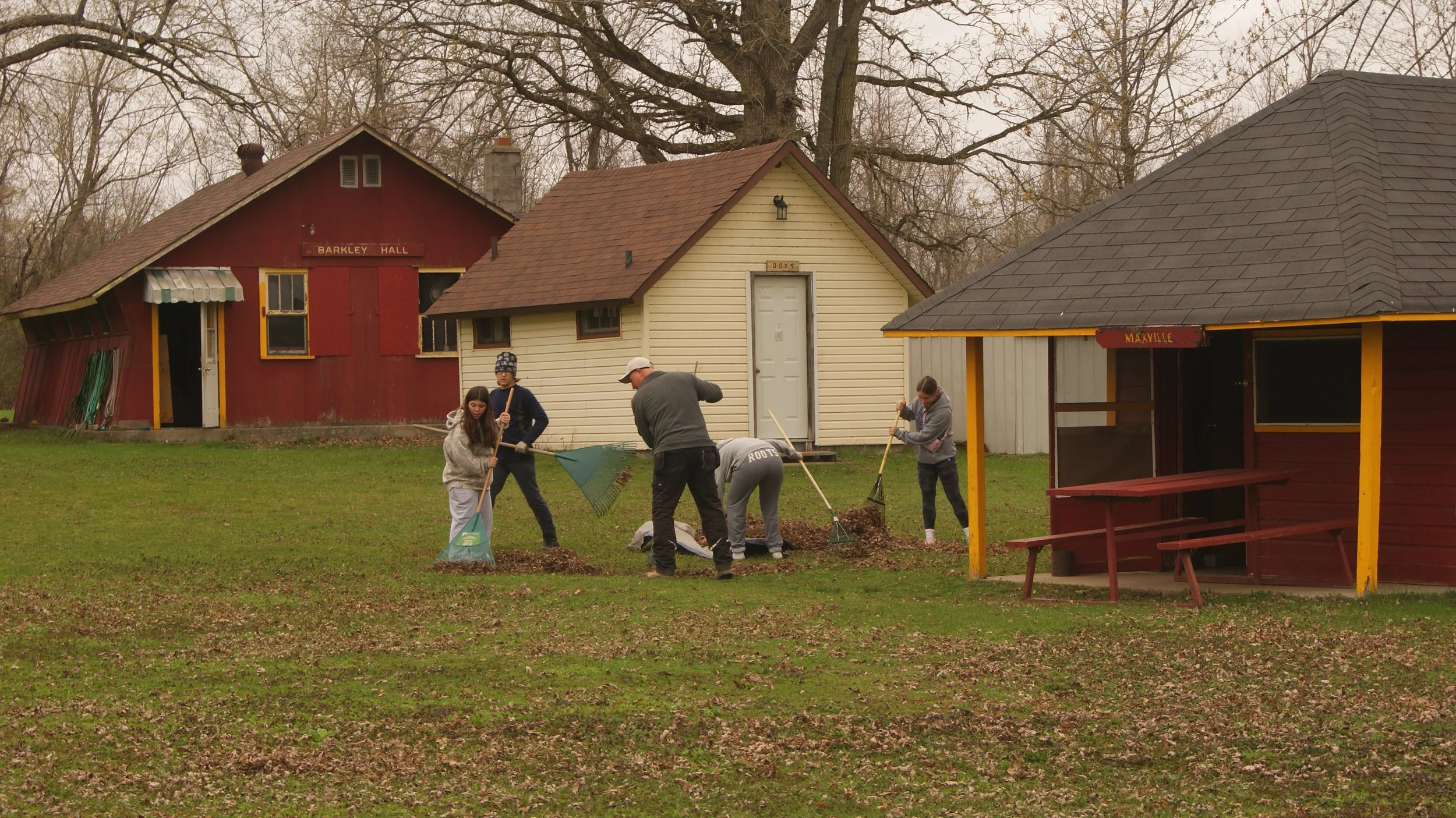 A group of people raking leaves in a grassy yard with small colored buildings in the background.