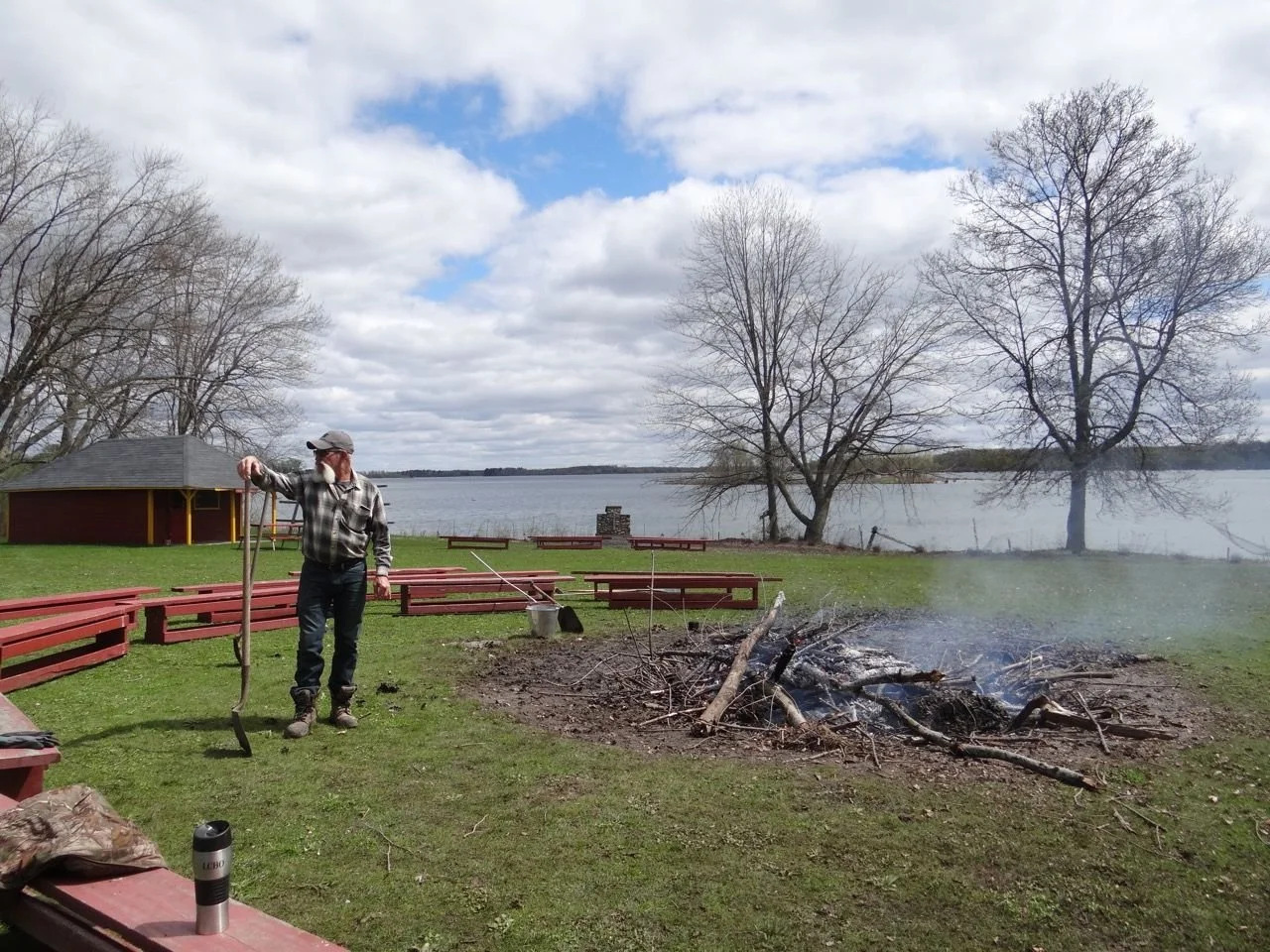 A man in a checkered shirt and baseball cap stands near a campfire with burning logs and sticks on a grassy lakeside park during daytime with cloudy skies and leafless trees.