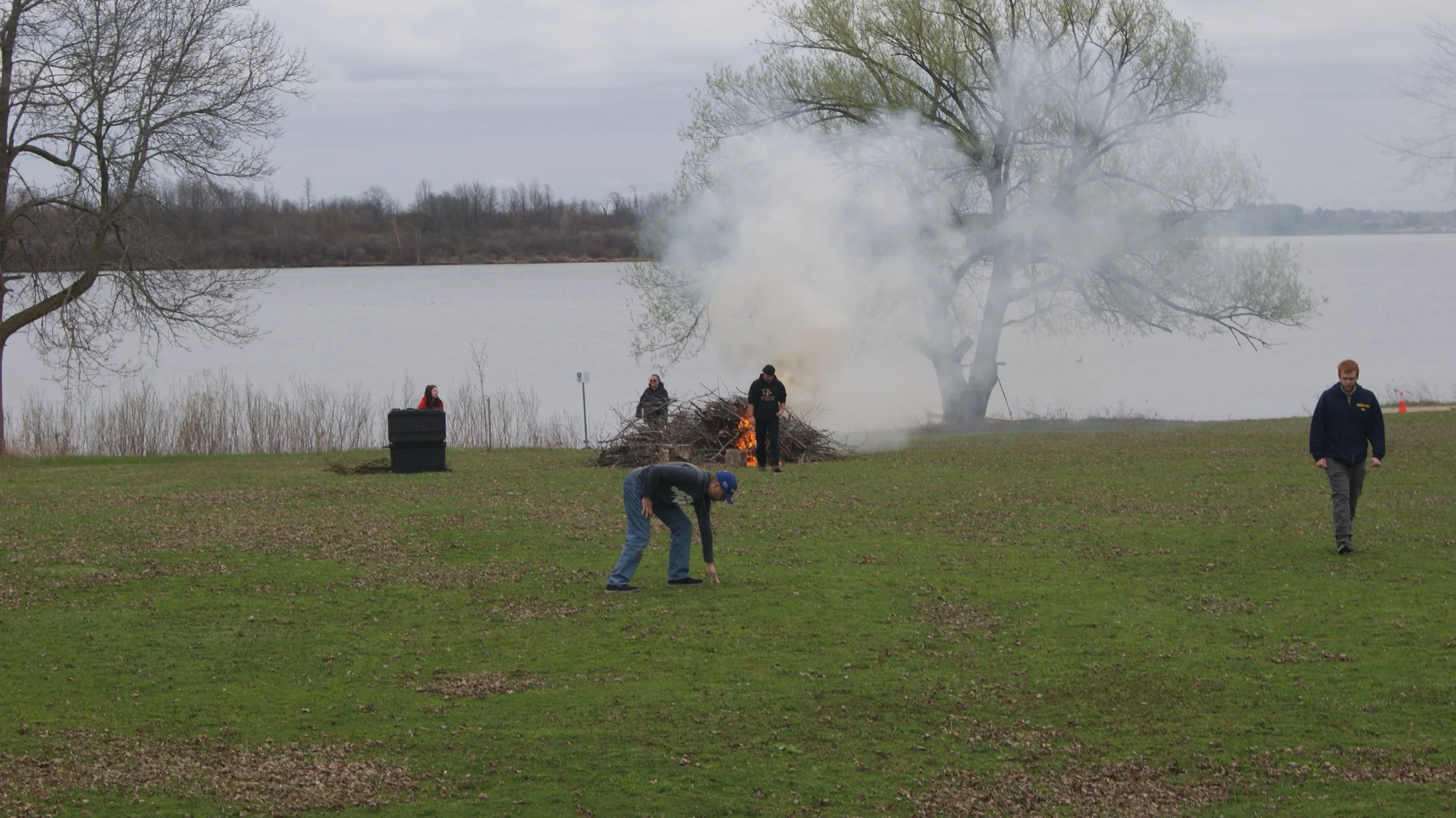 People burn a pile of sticks and branches near a lake with trees in the background.