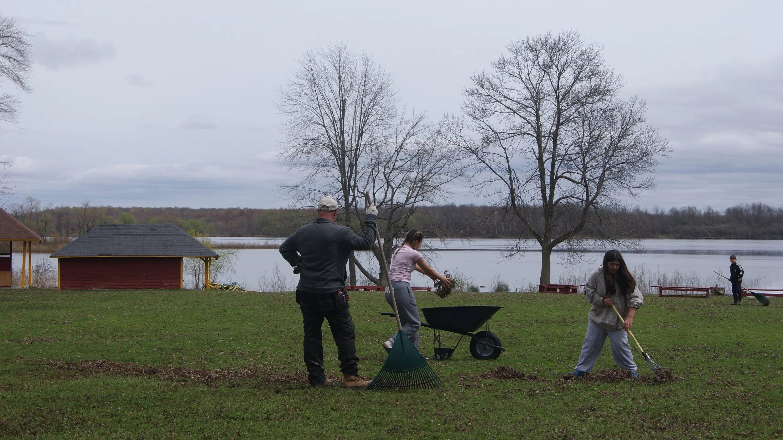 People raking leaves near a lake with leafless trees, a small red building, and an overcast sky in the background.