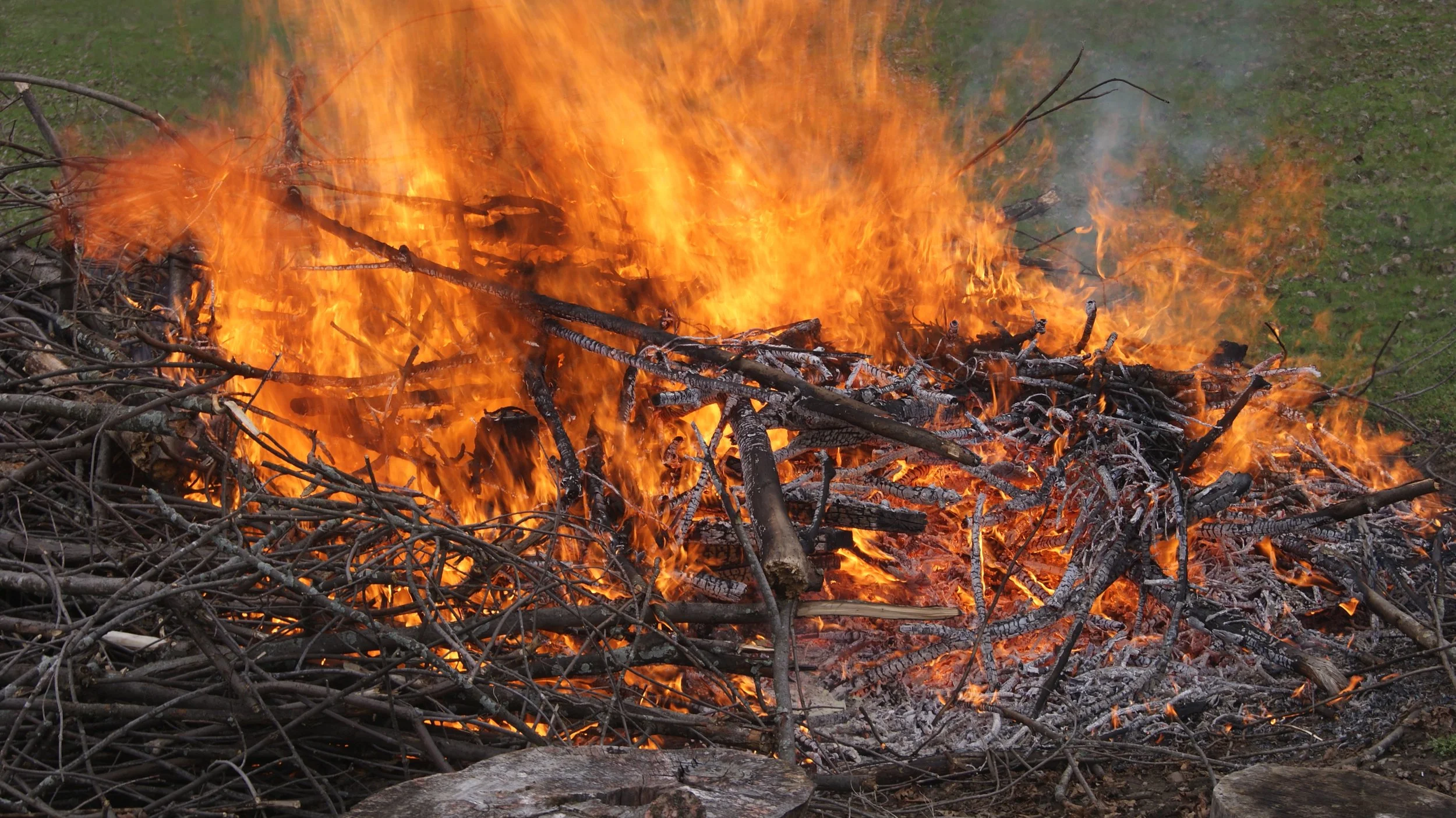 Burning pile of branches and logs with orange flames and smoke.