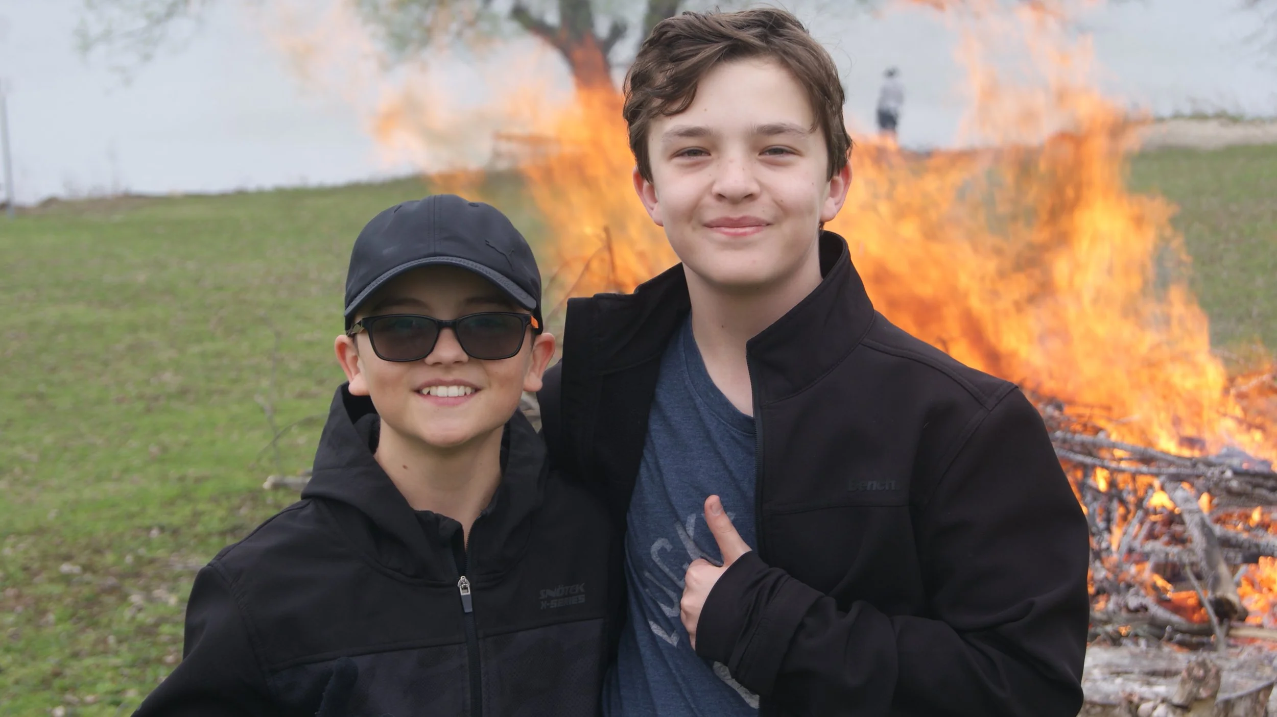 Two young boys standing outdoors in front of a fire. One boy wears a black cap and sunglasses, the other boy has curly hair and is smiling. They are smiling and wearing black jackets.