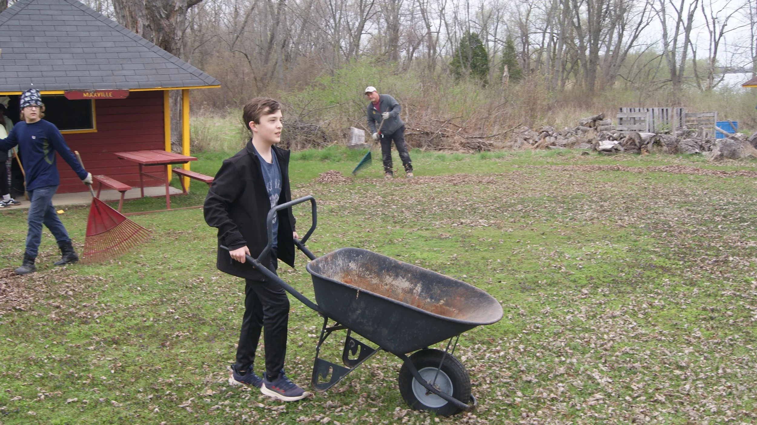A young boy in a black jacket and black pants is pushing a wheelbarrow across a grassy yard, with a red building and other children raking leaves in the background, during overcast weather.