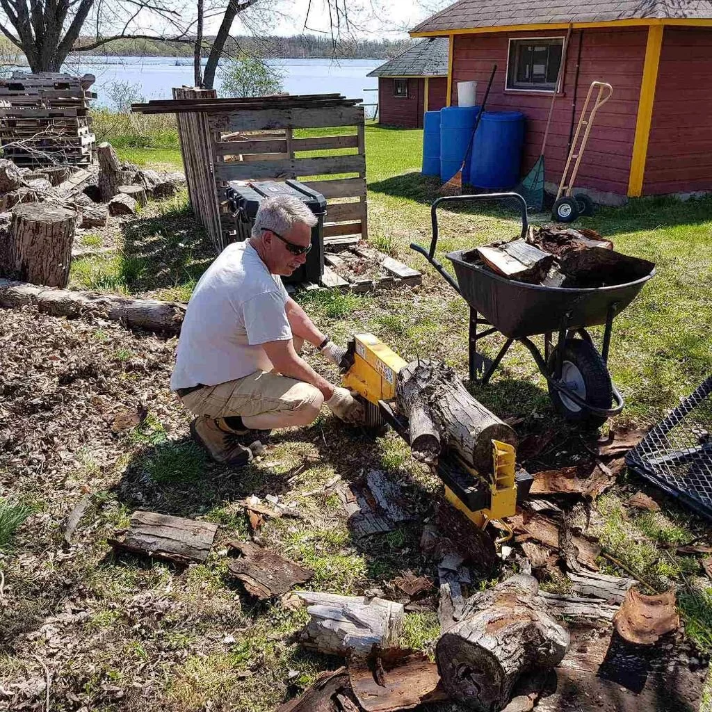 A man wearing sunglasses and khaki shorts is using a chainsaw to cut a large log into smaller pieces outside near a red shed with blue barrels. There are wood logs and chopped wood around him, and a wheelbarrow filled with wood pieces nearby. A lake 