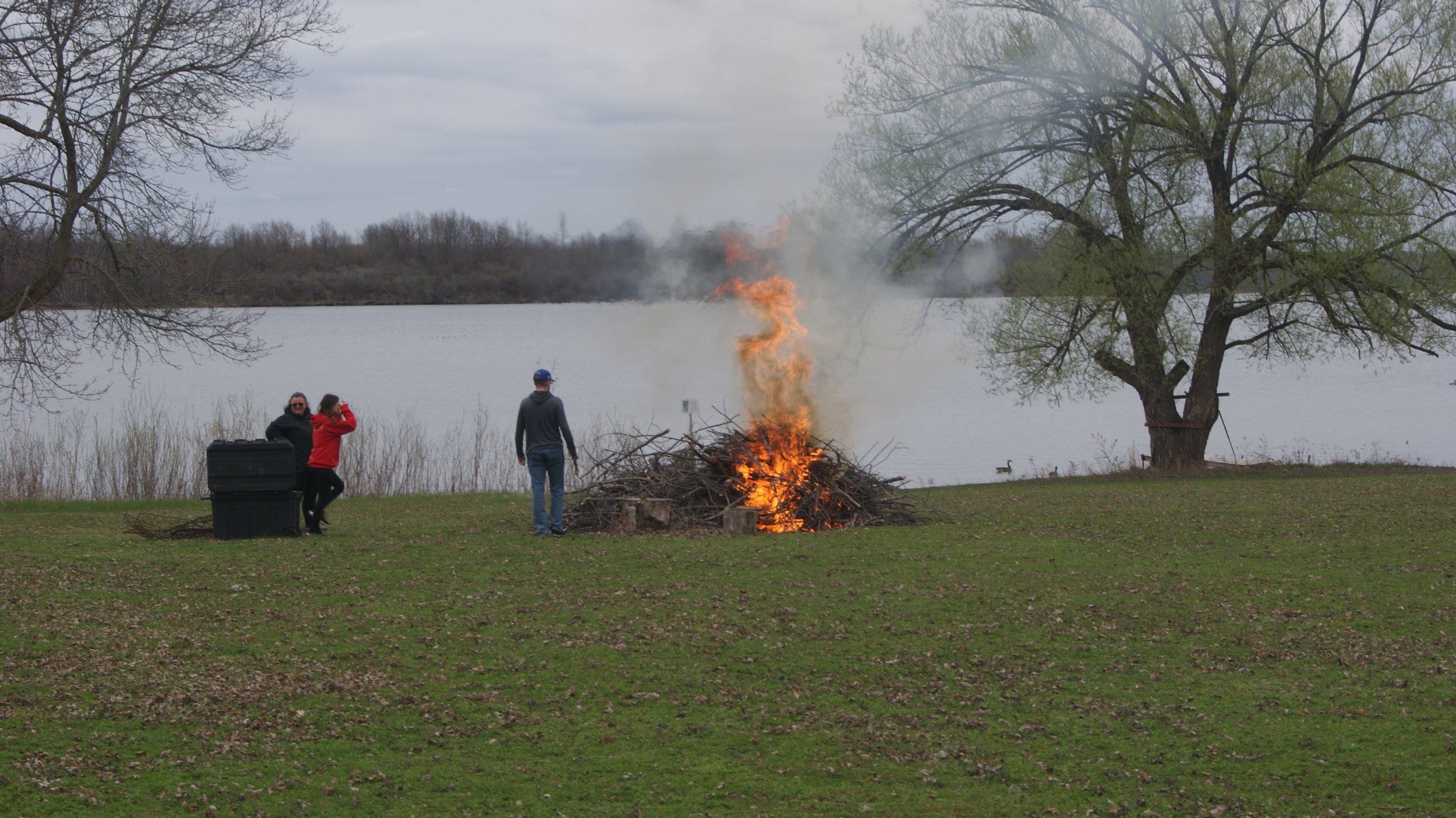 People near a campfire by a lake, with trees in the background and ducks swimming in the water.