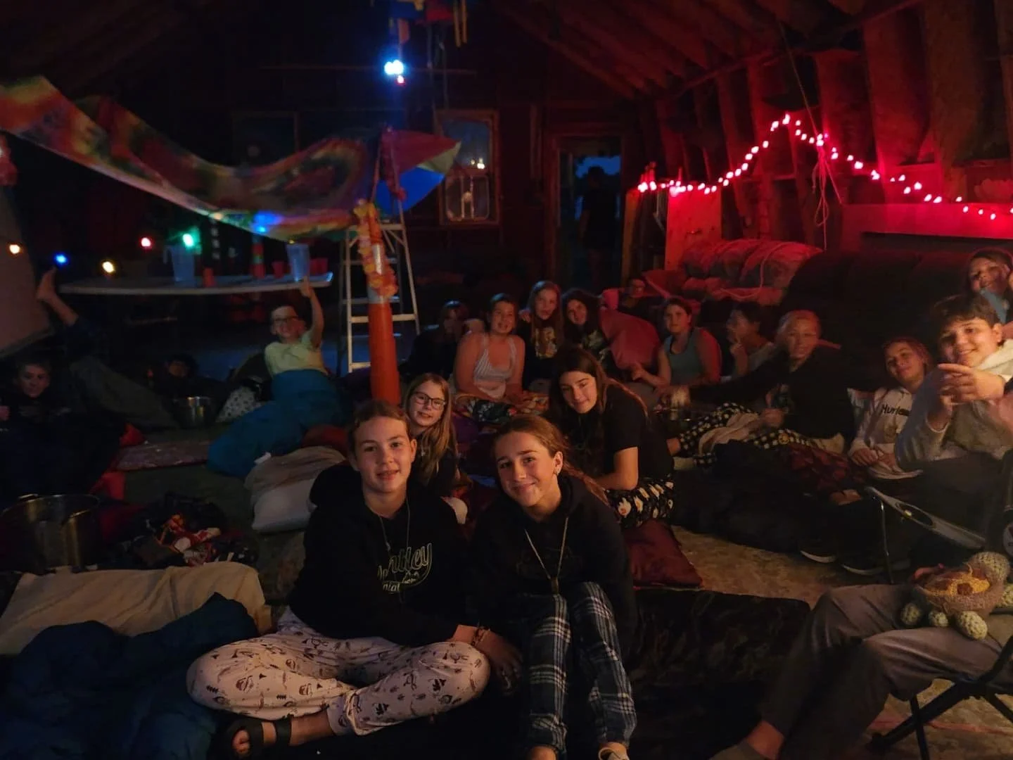A group of children sitting on the floor inside a decorated cabin at nighttime, with colorful string lights and fabric hanging from the ceiling, smiling and looking at the camera.