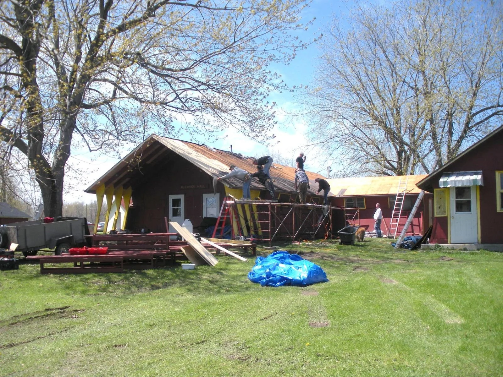 People working on a house roof renovation, with scaffolding and ladders, in a grassy yard with trees.