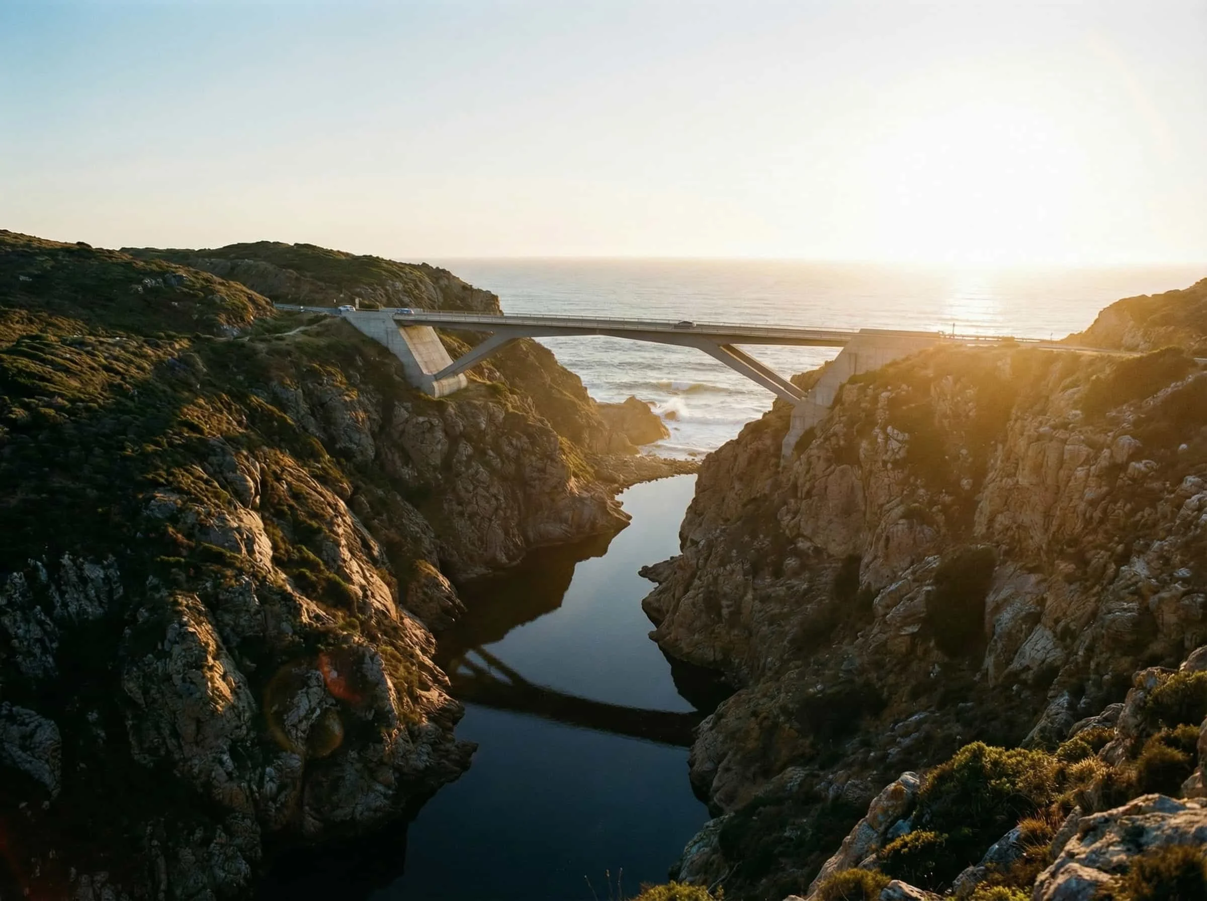 A bridge spanning a rocky canyon over a body of water with the ocean in the background, during sunset.