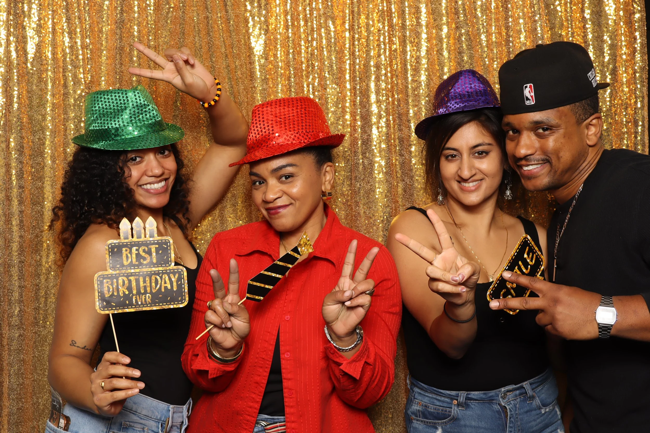 Group of four friends celebrating a birthday, wearing colorful hats and holding party signs and props, in front of a gold sequin backdrop.