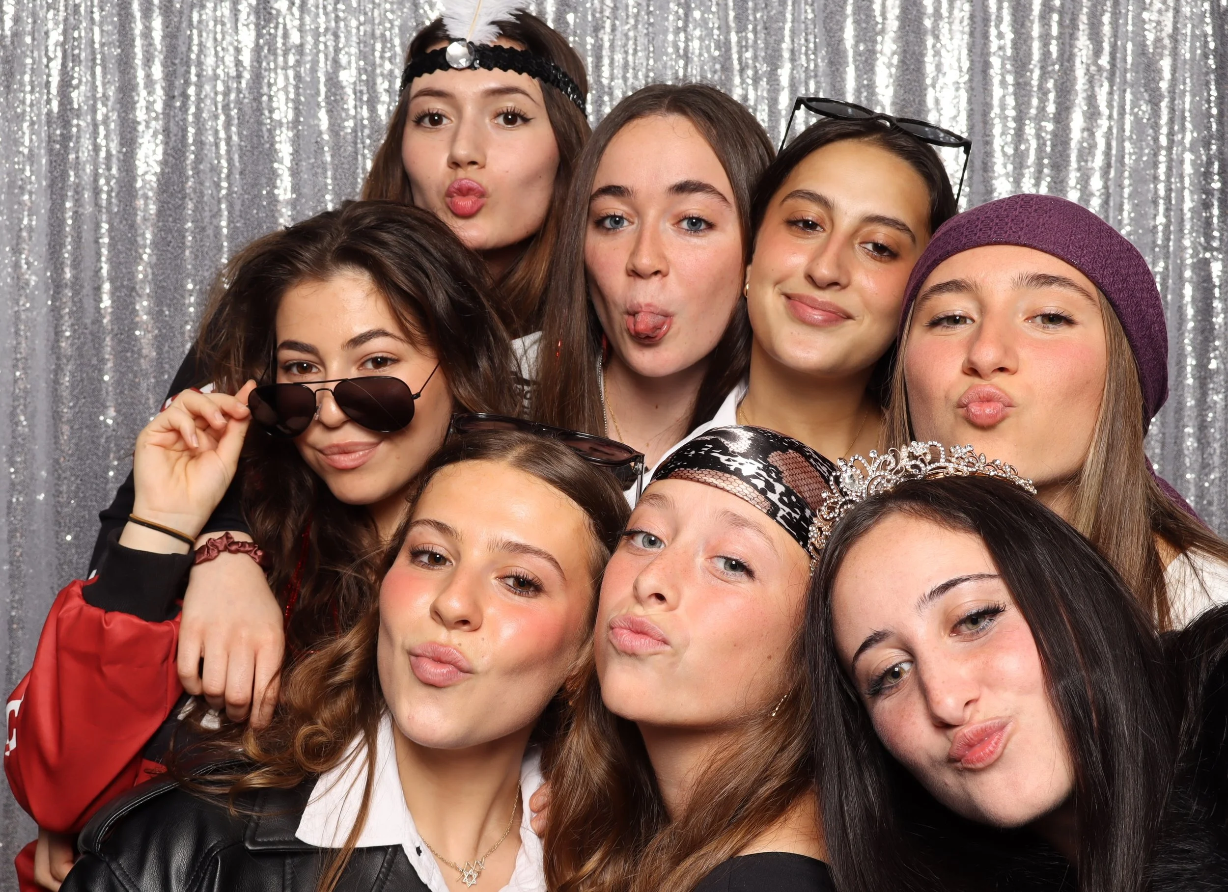 Group of nine young women posing in front of a sparkly silver backdrop, some wearing accessories like glasses, a tiara, headbands, and a beanie, making playful facial expressions.