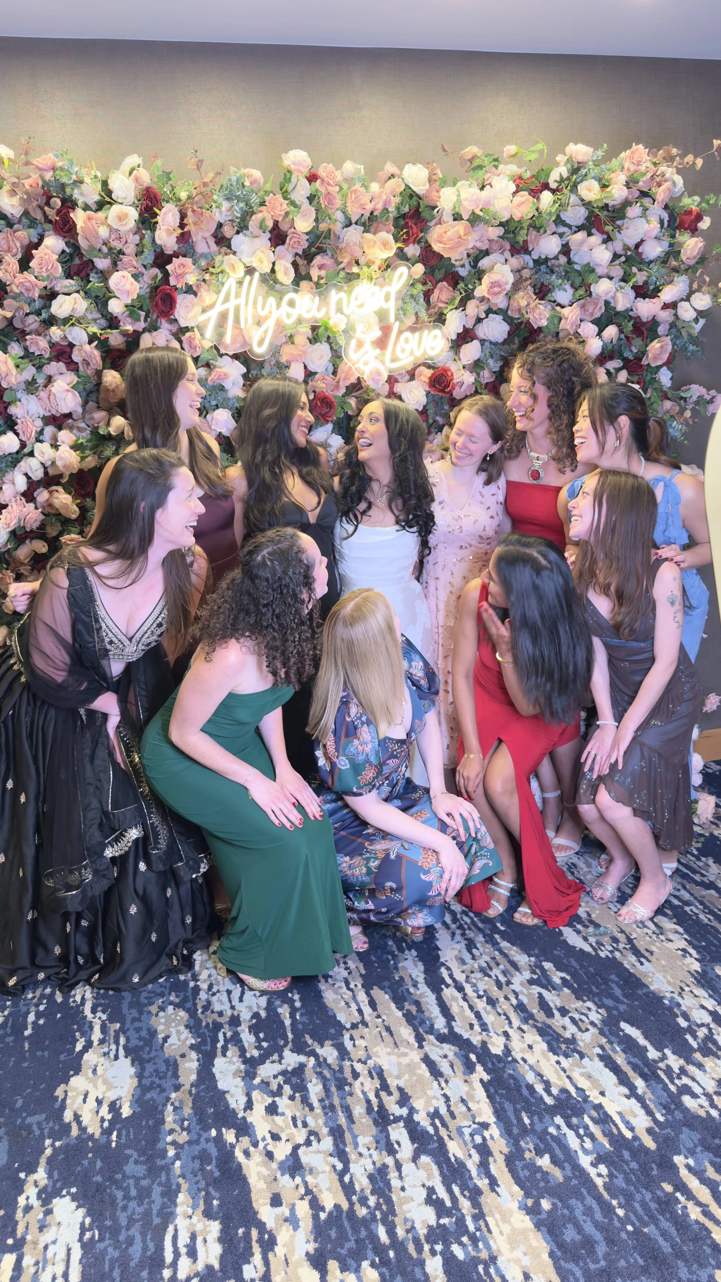 Group of women dressed in colorful dresses sharing a moment of joy in front of a floral backdrop with a neon sign that says 'All you need is love'.