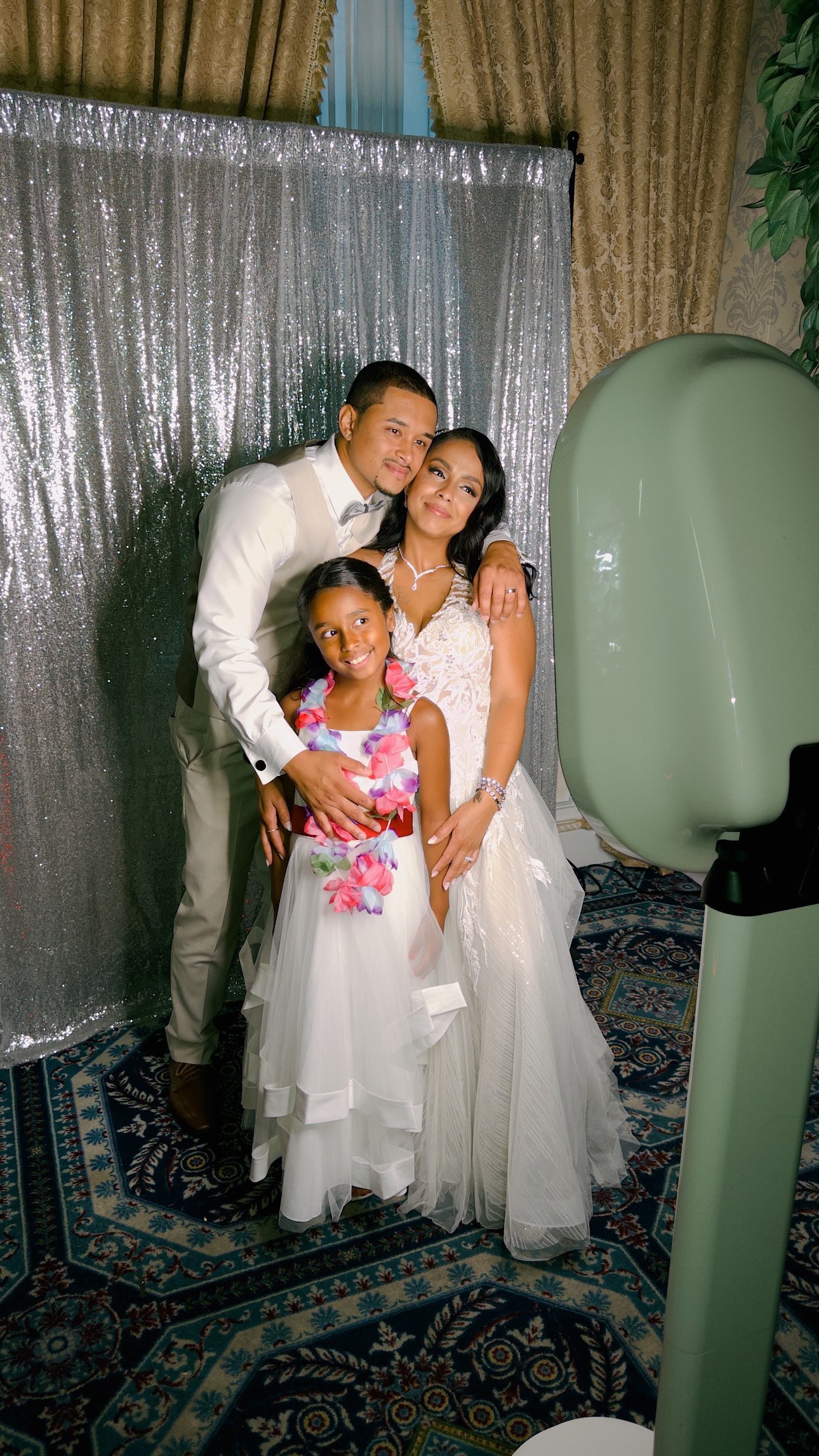 A family of three posing for a photo at a wedding reception, with a silver sequin backdrop and a photo booth prop machine nearby.