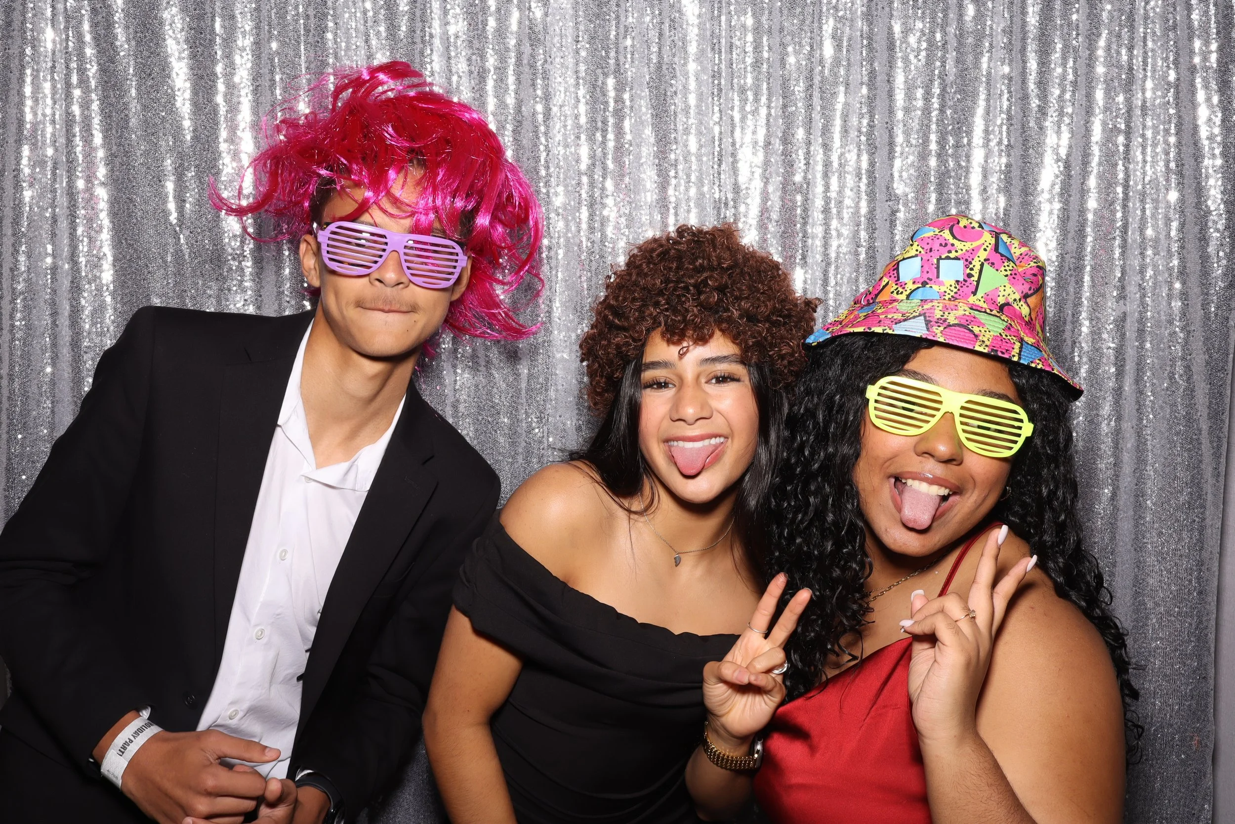 Three friends at a party wearing colorful wigs and fun sunglasses, posing playfully in front of a silver sequin backdrop.