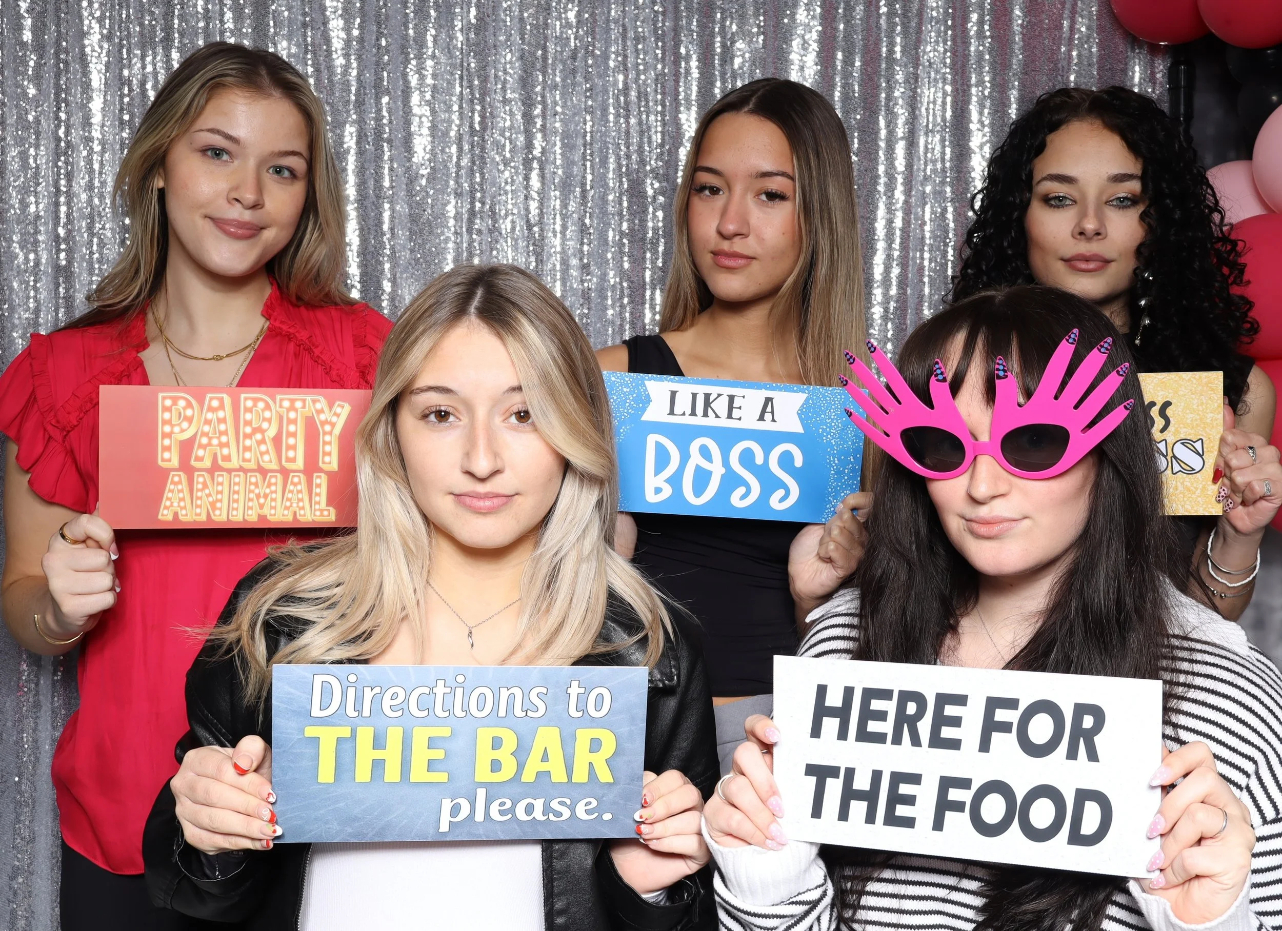 A group of five young women at a party holding humorous signs and props, with a silver sequin backdrop and pink, black, and red balloons in the background.